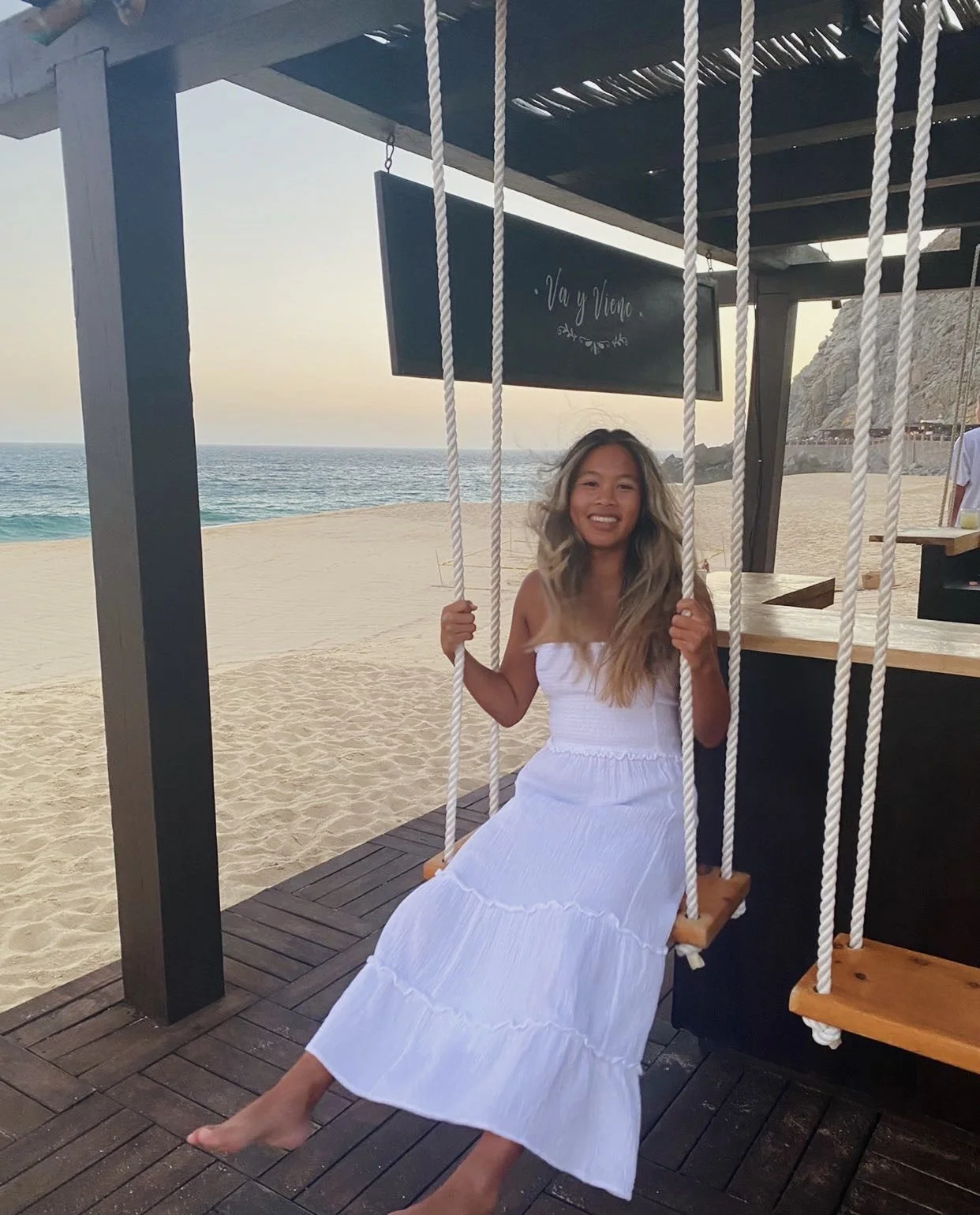 A woman in a white dress sitting on a swing at the beach, smiling, with the ocean and sky in the background and a sign that says 'Vaj y Viene' above her.
