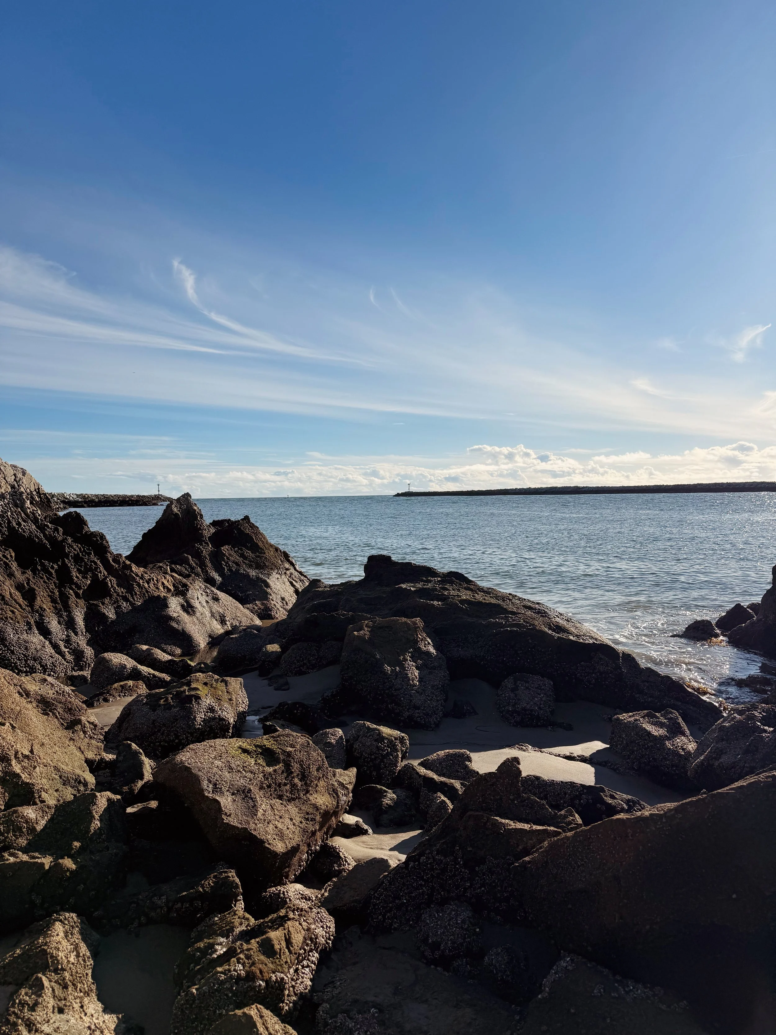 Rocky shoreline with large rocks and calm water under a blue sky with wispy clouds.