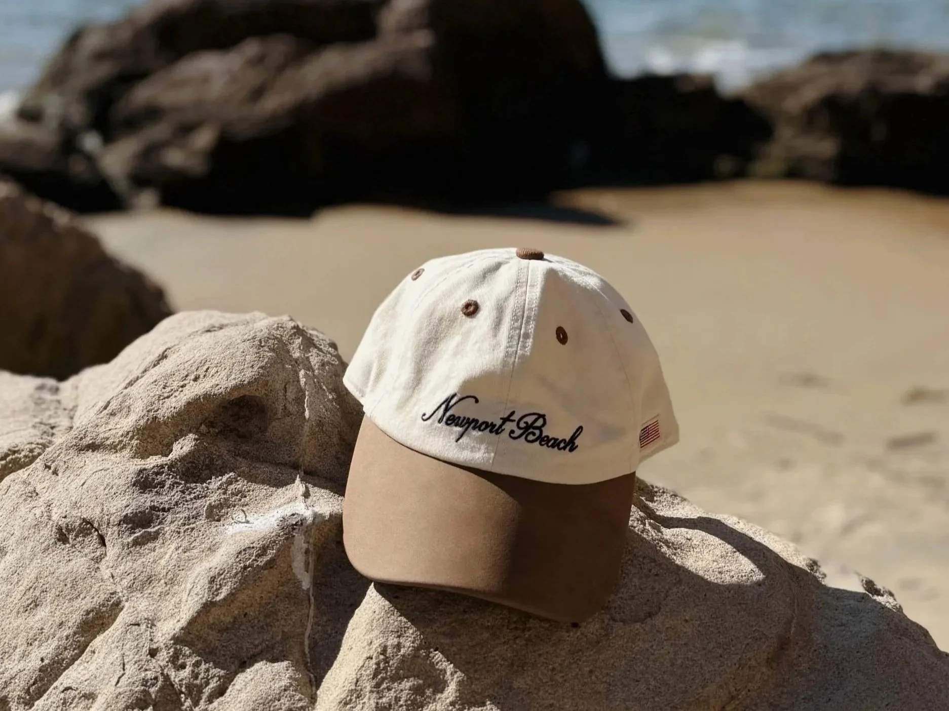 A baseball cap with a white top and a beige brim, embroidered with 'Newport Beach' in black and a small American flag patch, placed on a large rock at the beach.