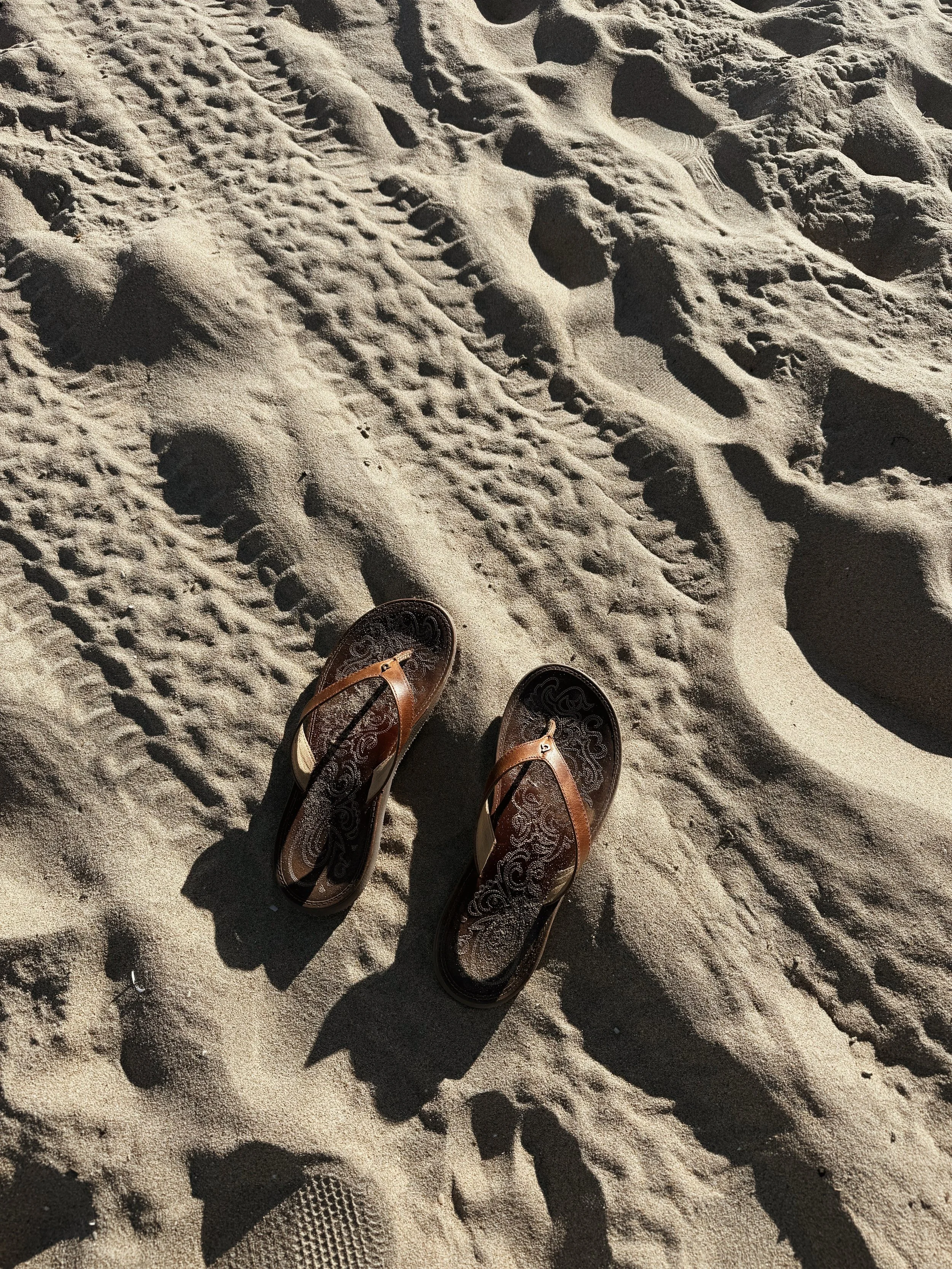 Brown flip-flops on the sandy beach with visible footprints and tire tracks in the sand.