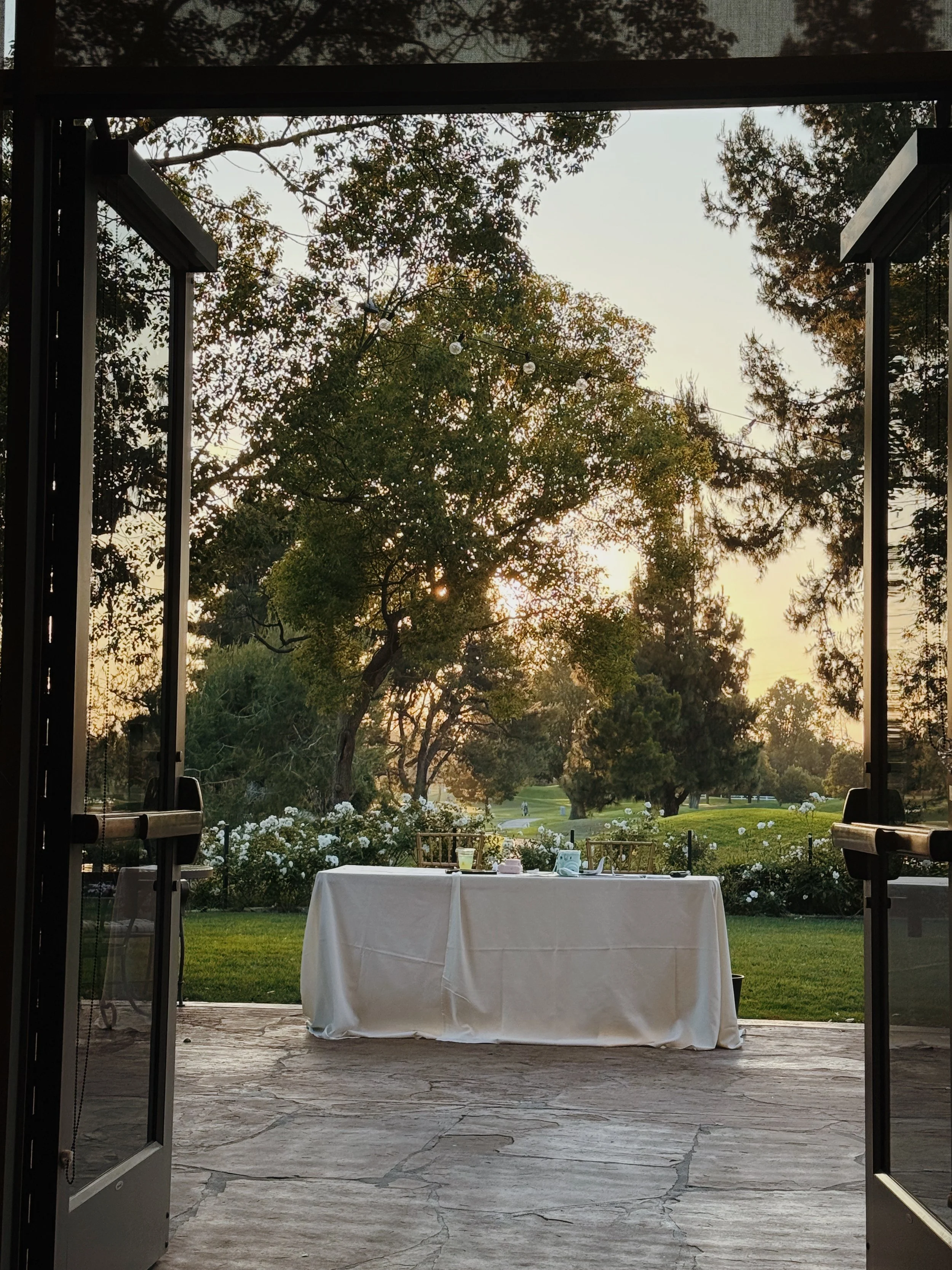 A table set for an outdoor dinner with a white tablecloth, glasses, and plates, viewed from open sliding glass doors, overlooking a garden with trees and white flowers at sunset.