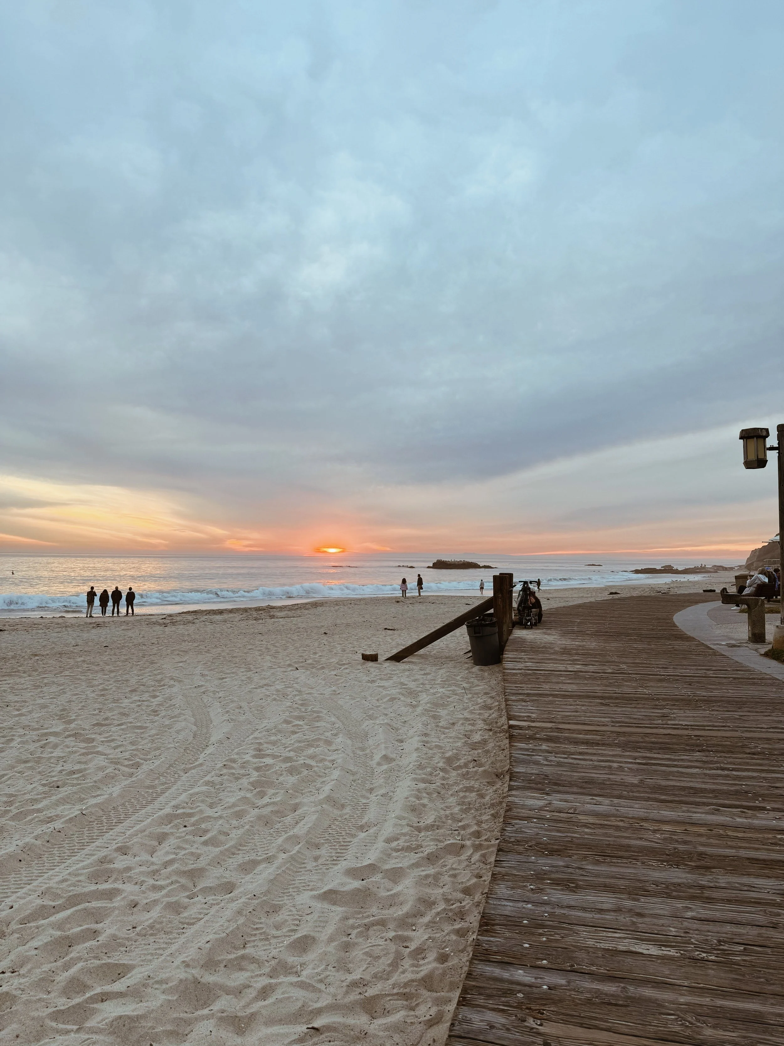 Beach at sunset with a wooden boardwalk, a few people standing near the water, and others sitting on benches. Cloudy sky with a hint of sunlight on the horizon.