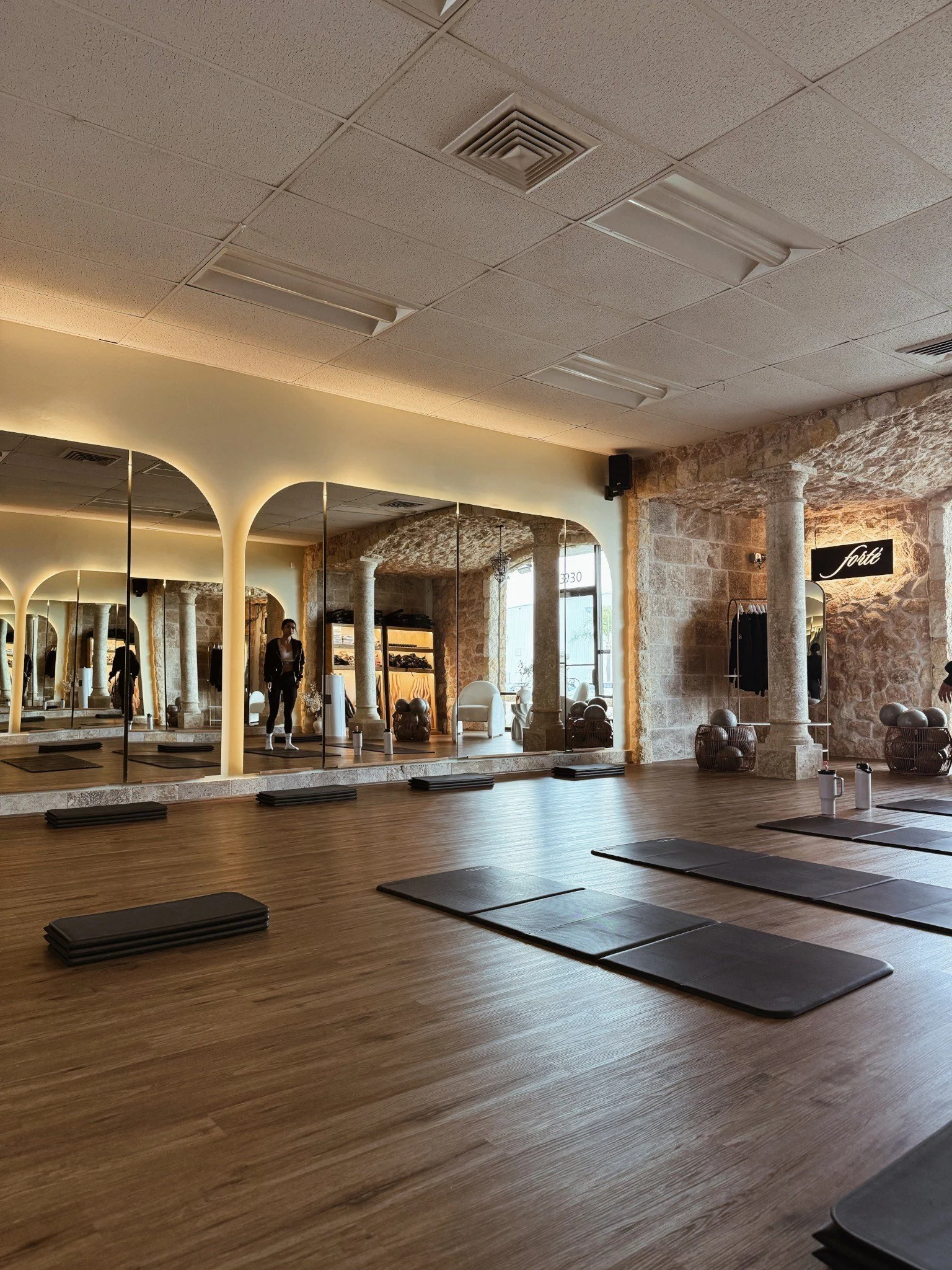 Empty yoga or fitness studio with wooden floors, black mats and water bottles, mirrored wall, stone columns, and modern lighting.