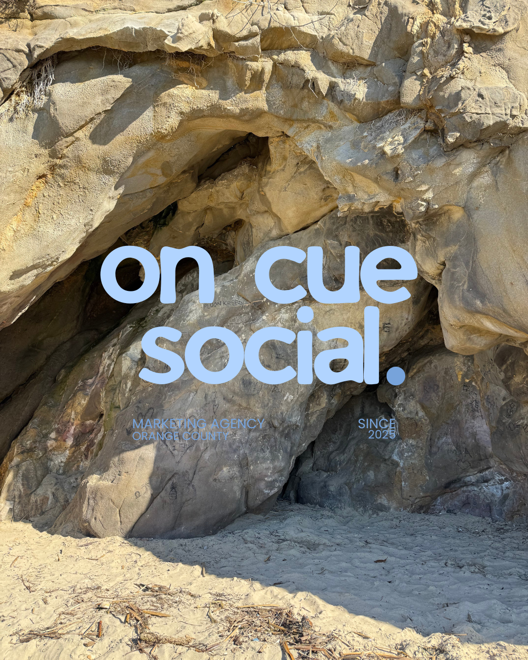 A rocky natural formation with large boulders on a sandy beach, overlaid with blue text that says 'on cue social.' and smaller text 'MARKETING AGENCY ORANGE COUNTY SINCE 2025'.