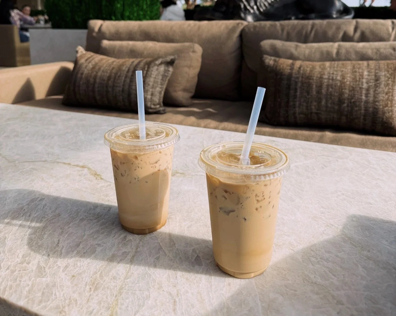 Two iced coffee drinks with straws on a white marble table in front of a beige couch with multiple brown cushions at an outdoor seating area.
