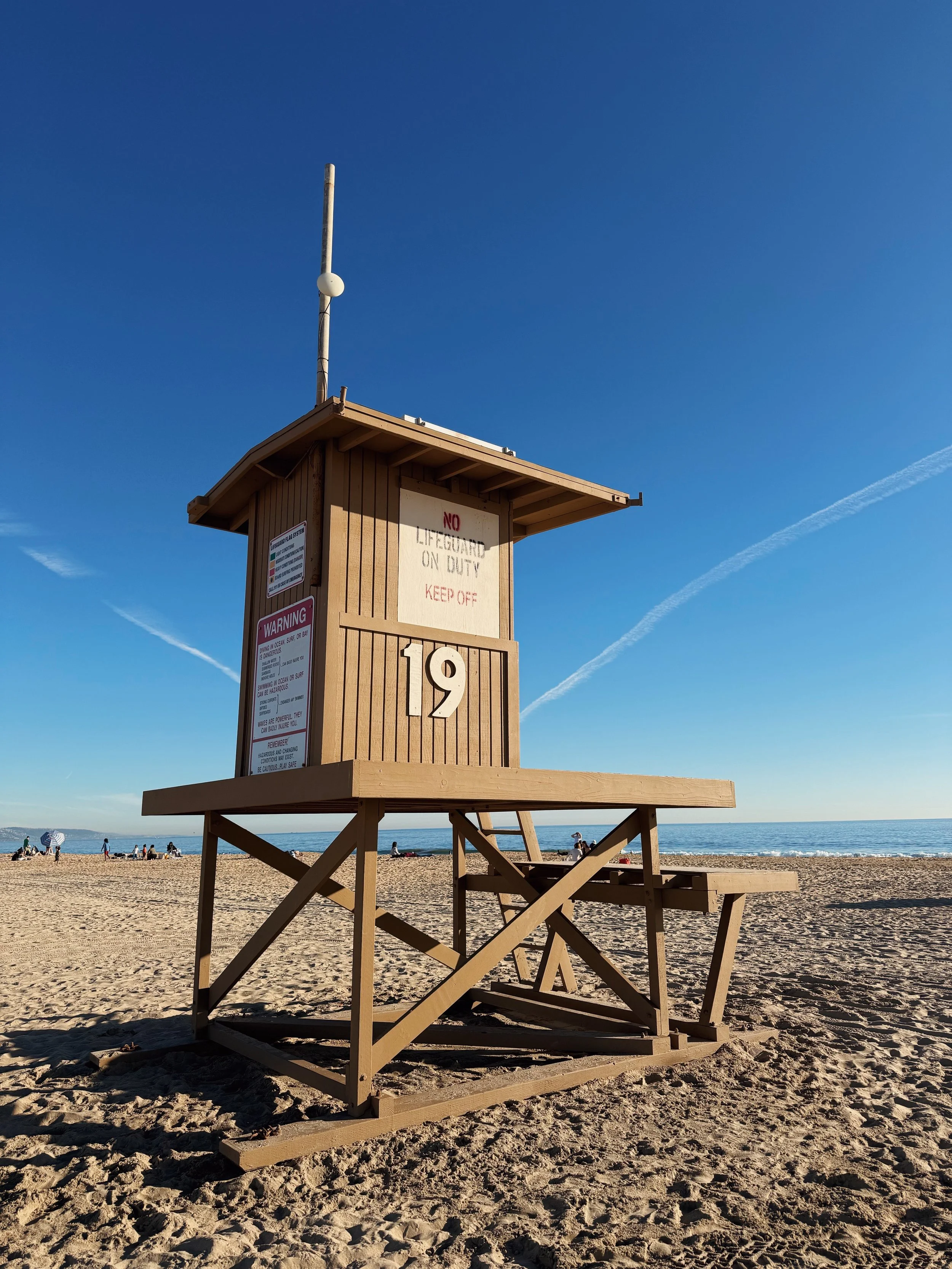 A wooden lifeguard tower numbered 19 on a sandy beach with a clear blue sky and ocean in the background.
