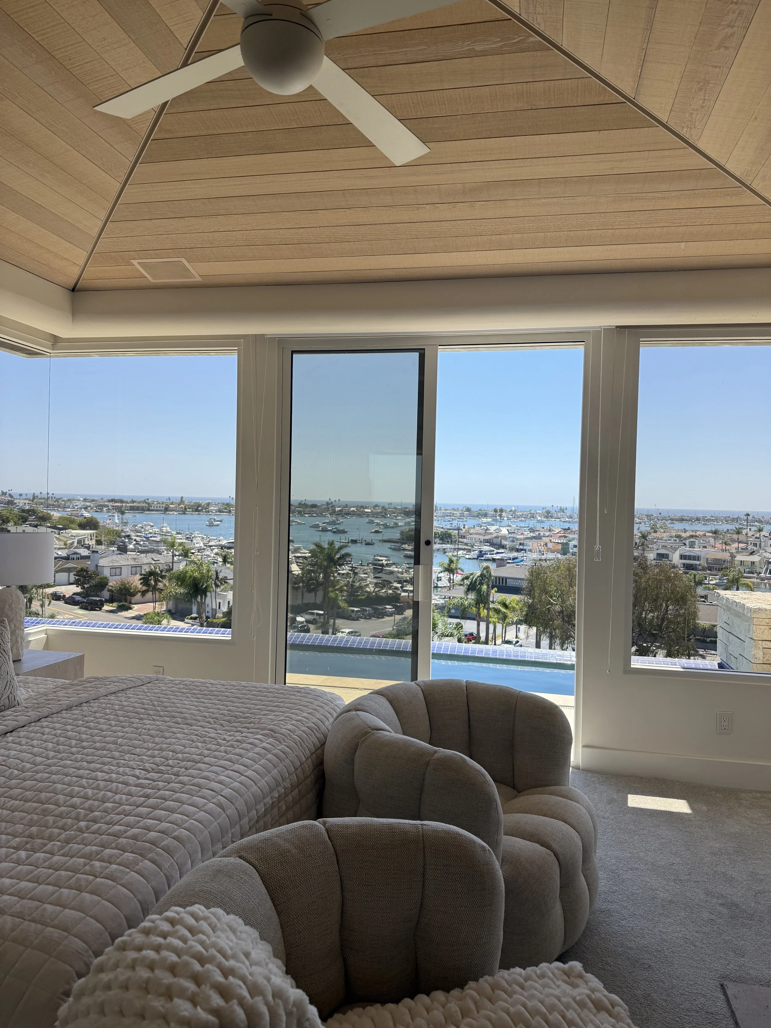 Living room with large windows overlooking a marina with boats, a cloudy sky, a beige chair, and a bed with a beige quilted blanket. The ceiling has wood paneling with a white ceiling fan.