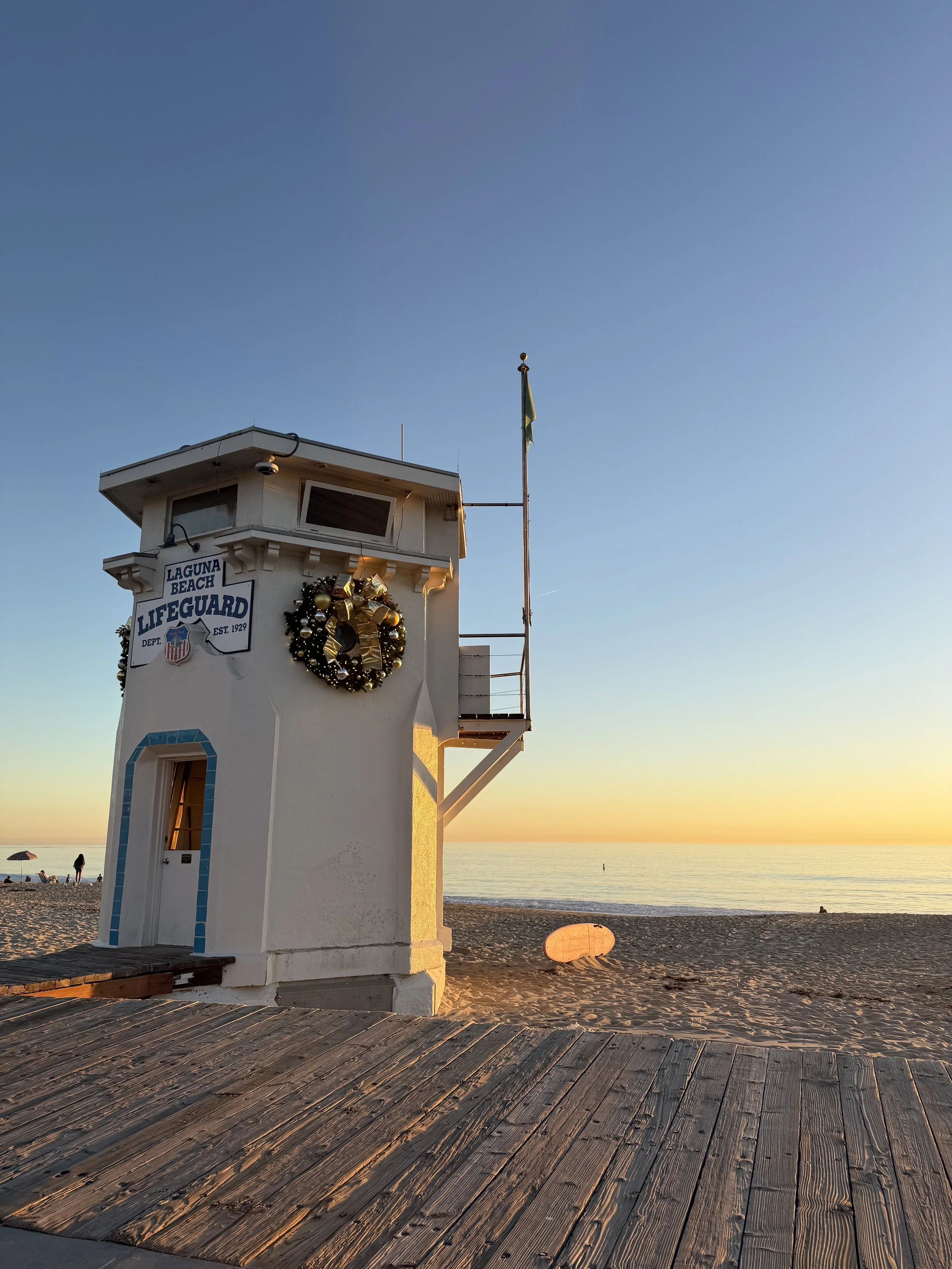 A white lifeguard tower on the beach decorated with a Christmas wreath, with the ocean and a sunset in the background.