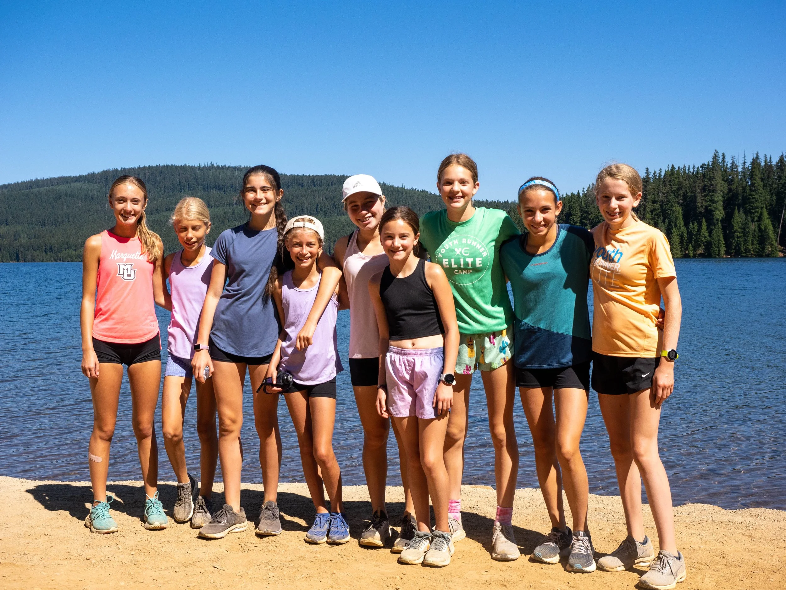Group of ten young girls standing on a sandy lakeshore, smiling, with a lake and forested hills in the background on a sunny day. Elite Middle School Camp at Youth Runner Camps