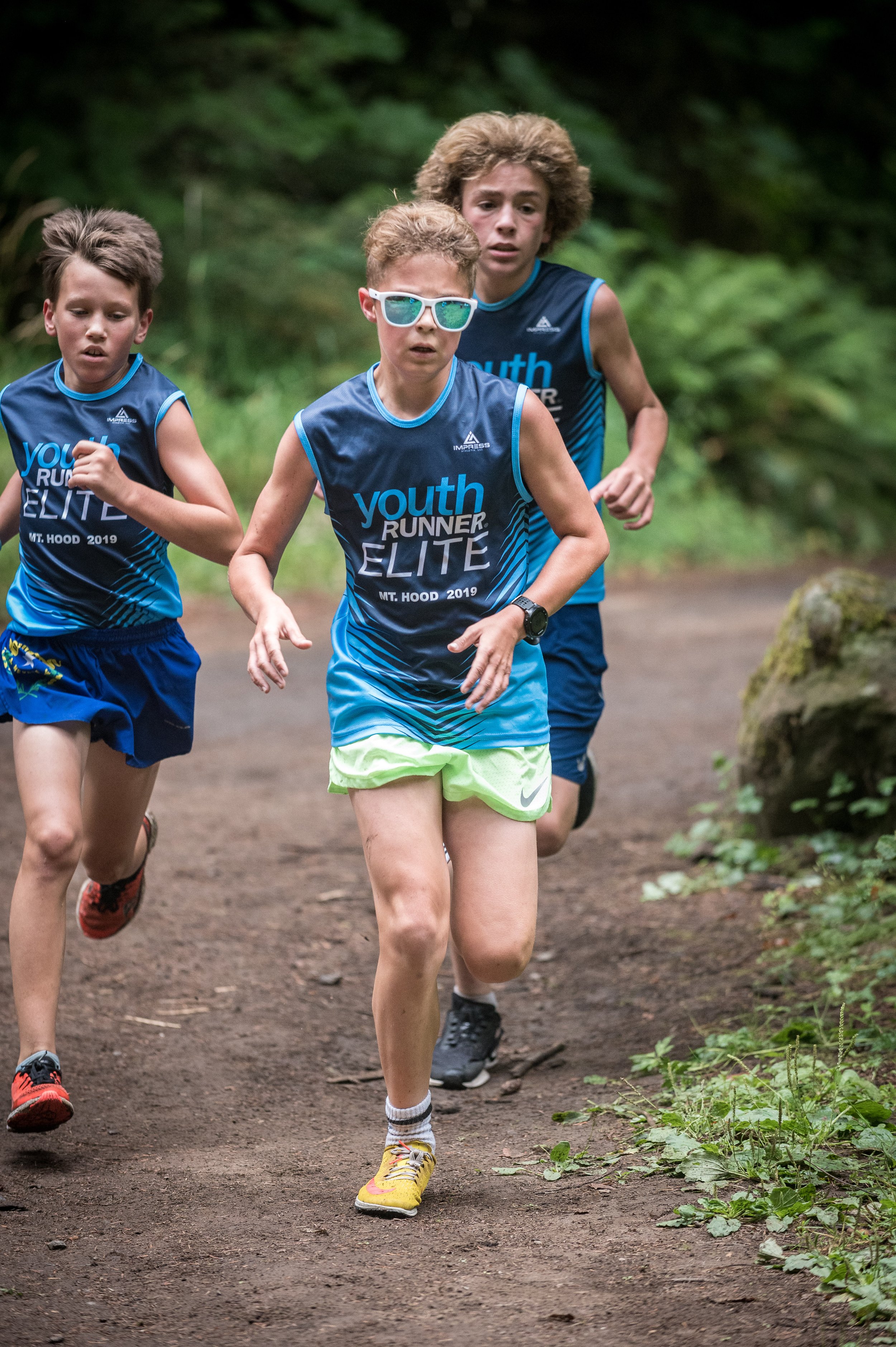 Three young boys running on a dirt trail in a wooded area, wearing blue athletic shirts that say 'youth RUNNER ELITE' and shorts.