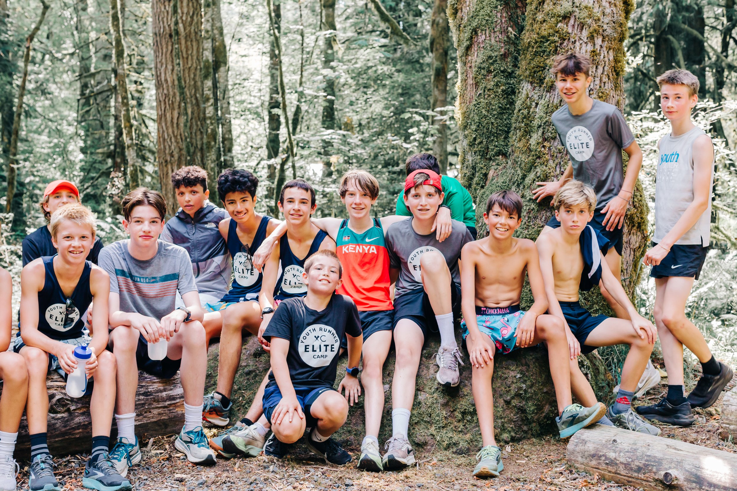 A group of 14 children, mostly shirtless or in casual athletic clothes, gathered around a large tree in a forest, smiling and posing for a photo at the Elite Middle School Camp at Youth Runner Camps