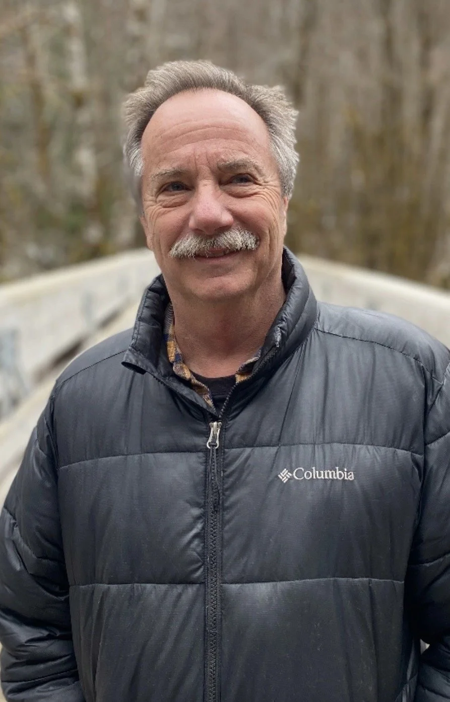 An older man with gray hair and a mustache smiling outdoors, wearing a black Columbia puffer jacket with a checkered shirt underneath, on a bridge with a blurry background of trees.