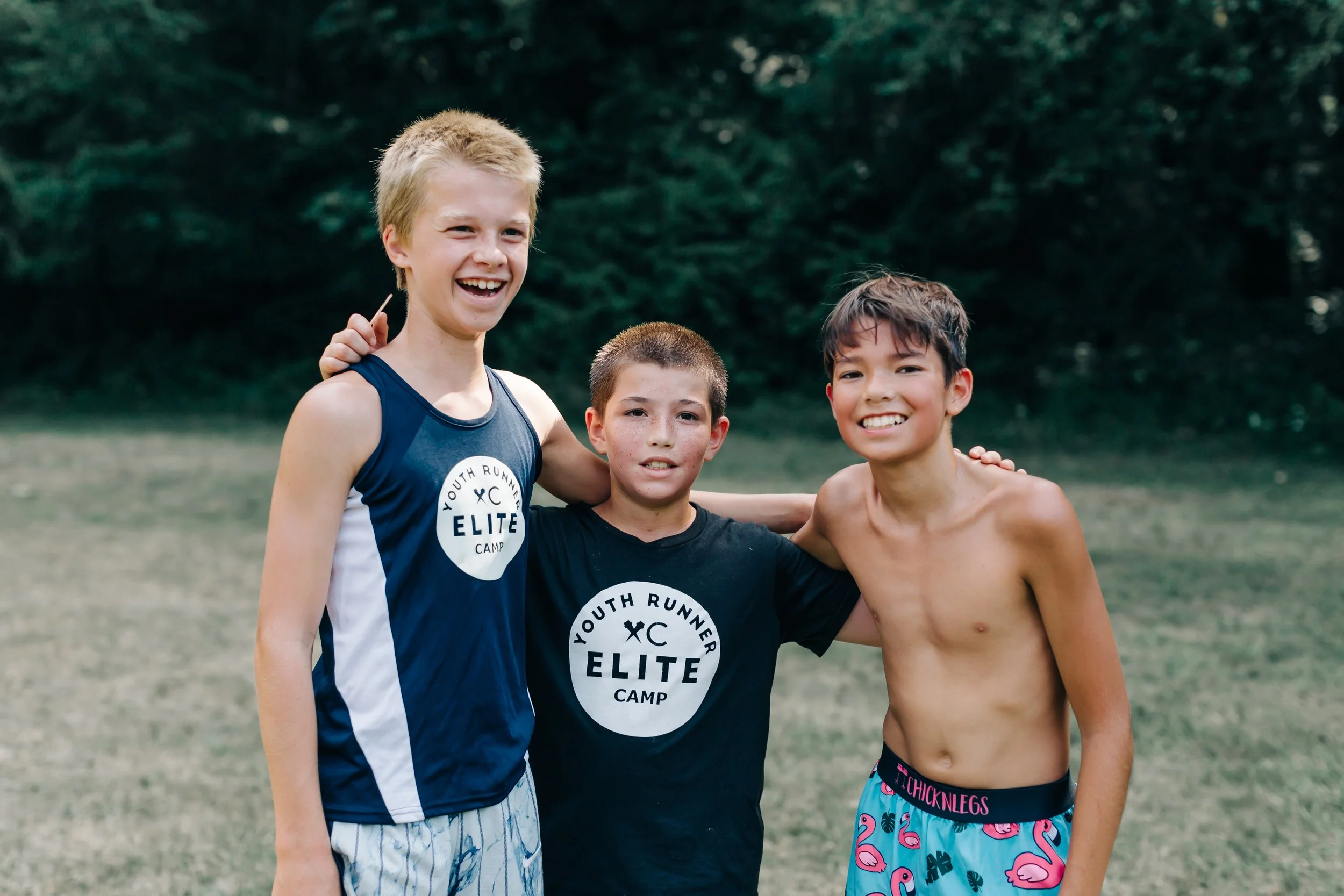 Three boys standing outdoors on grass, smiling and with arms around each other, two wearing black t-shirts with "Youth Runner Elite Camp" logo, one shirtless wearing colorful swim shorts with flamingos.