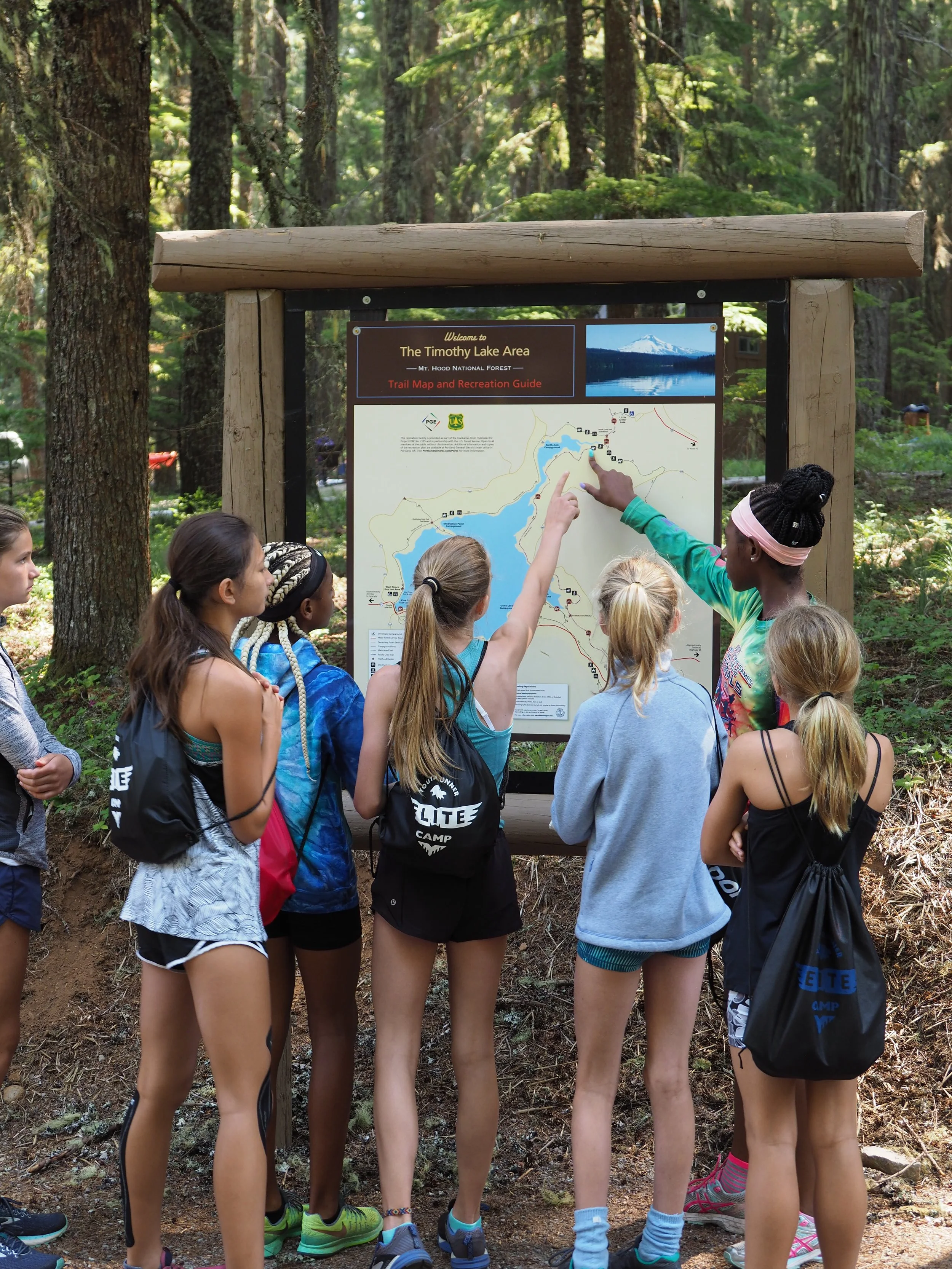 A group of young girls in athletic clothing and backpacks study a trail map at Timothy Lake area in a forest, with a park ranger or instructor pointing at the map at the Elite Middle School Camp at Youth Runner Camps