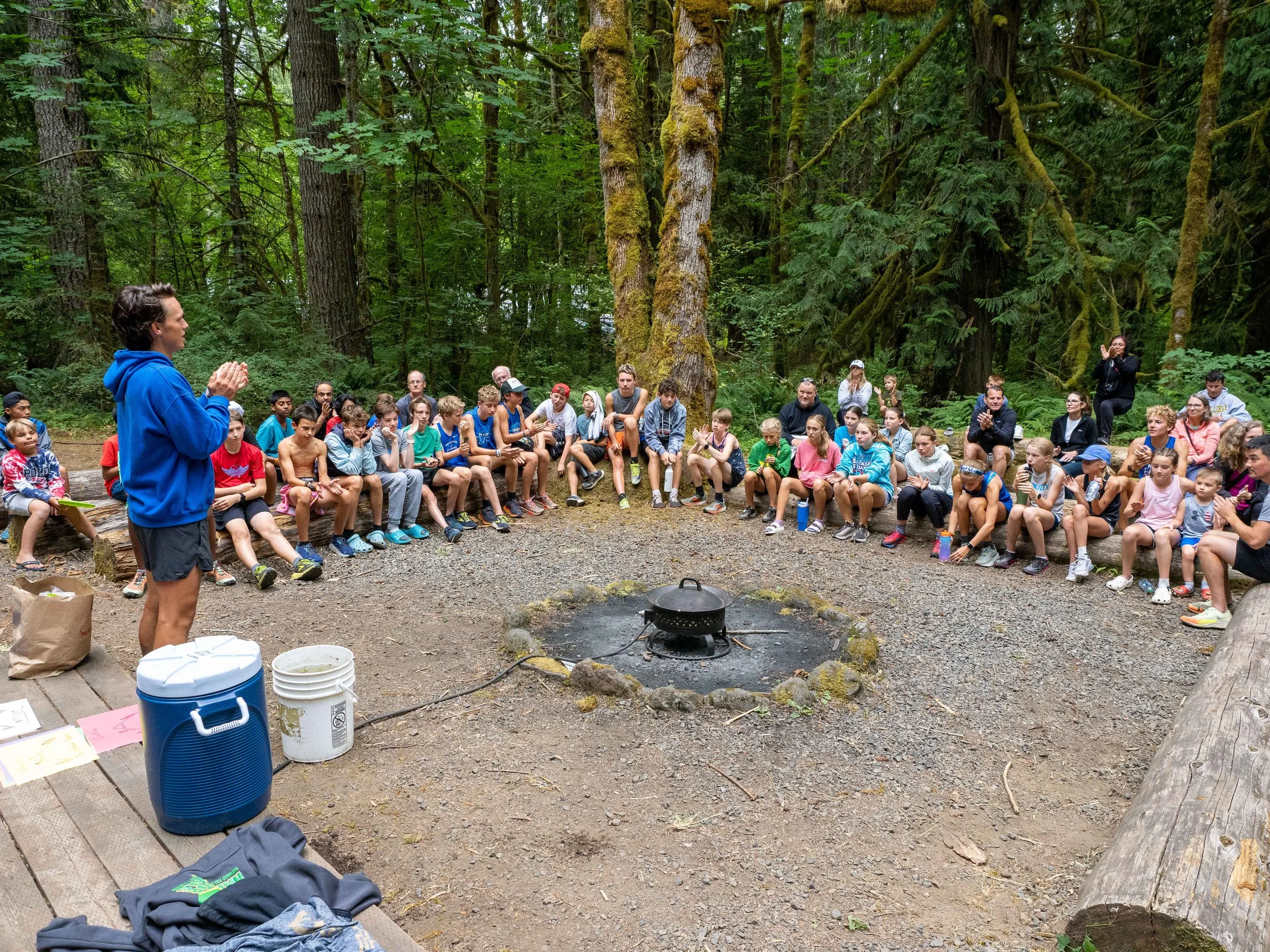 Group of young athletes and adults sitting in a circle around a campfire in a forest during daytime, listening to a person standing and speaking. The Elite Middle School Camp at Youth Runner Camps