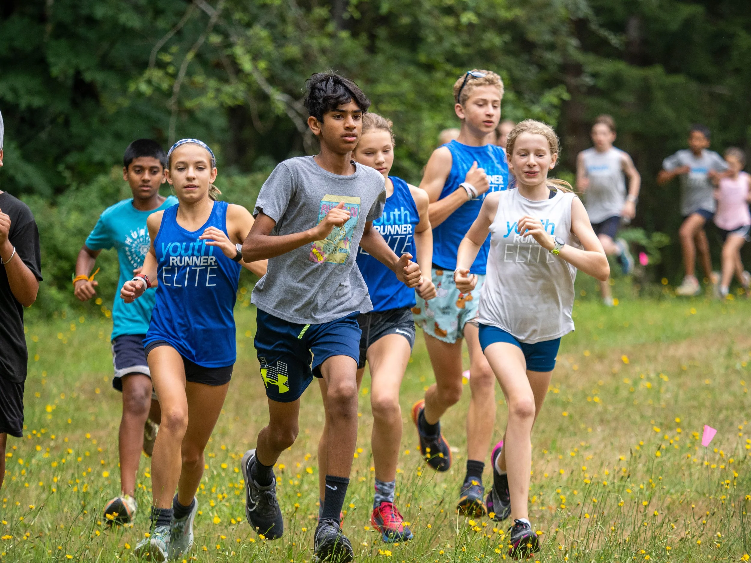 Young athletes running together in a grassy outdoor area during a race or running event. The Elite Middle School Camp at Youth Runner Camps