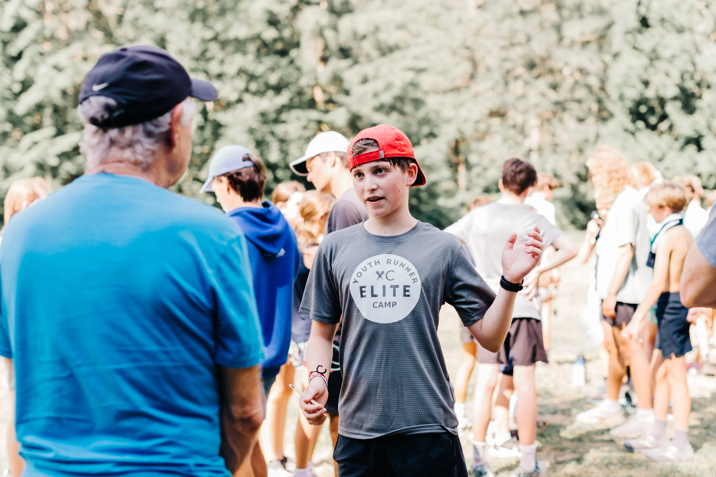 A young boy in a gray shirt and red cap engaged in conversation with an older man in a blue shirt and black cap during an outdoor running event, with several other participants in the background at the Elite Middle School Camp at Youth Runner Camps