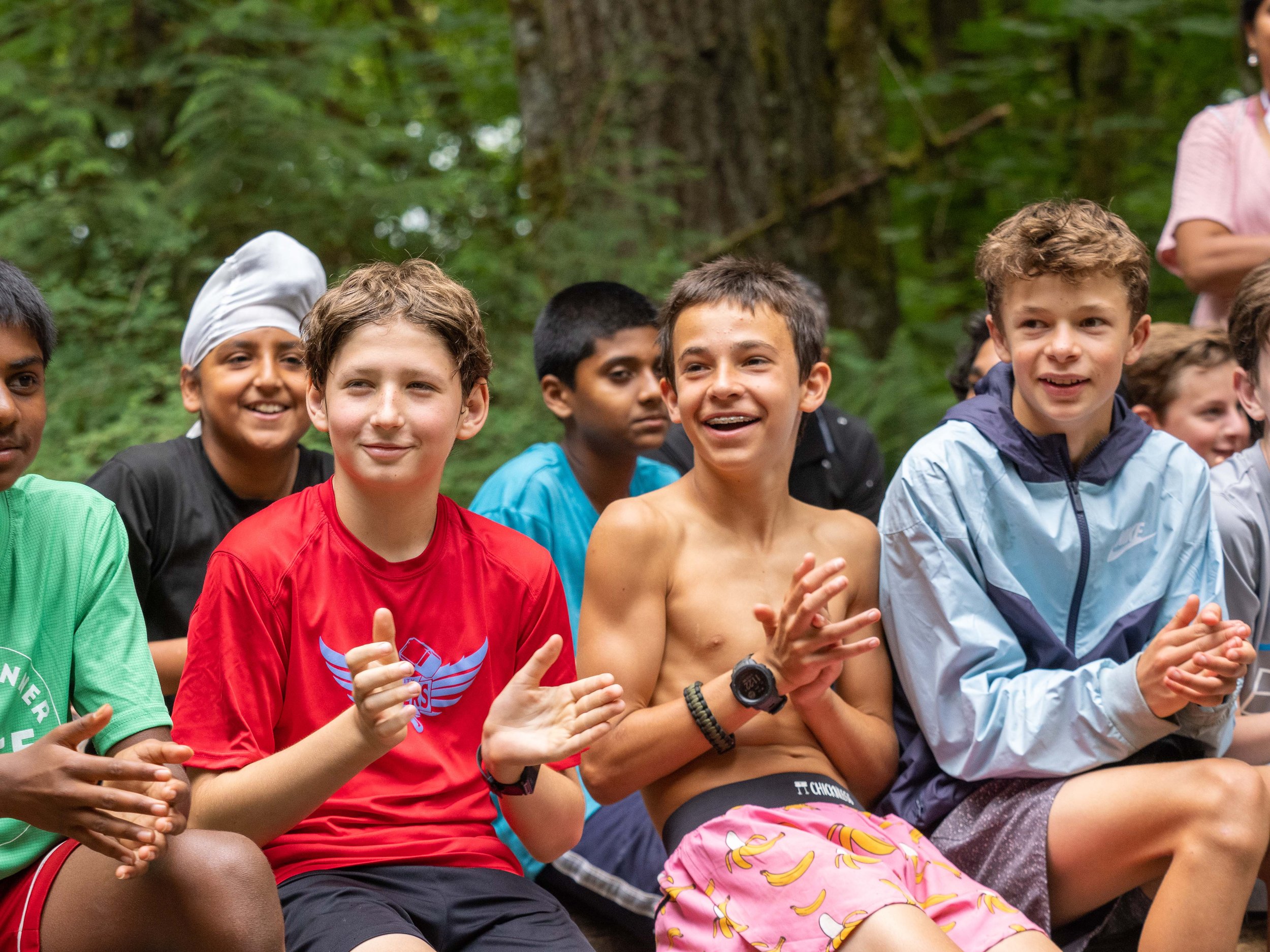 A group of smiling boys sitting outdoors in a wooded area, clapping and enjoying themselves at the Elite Middle School Camp at Youth Runner Camps
