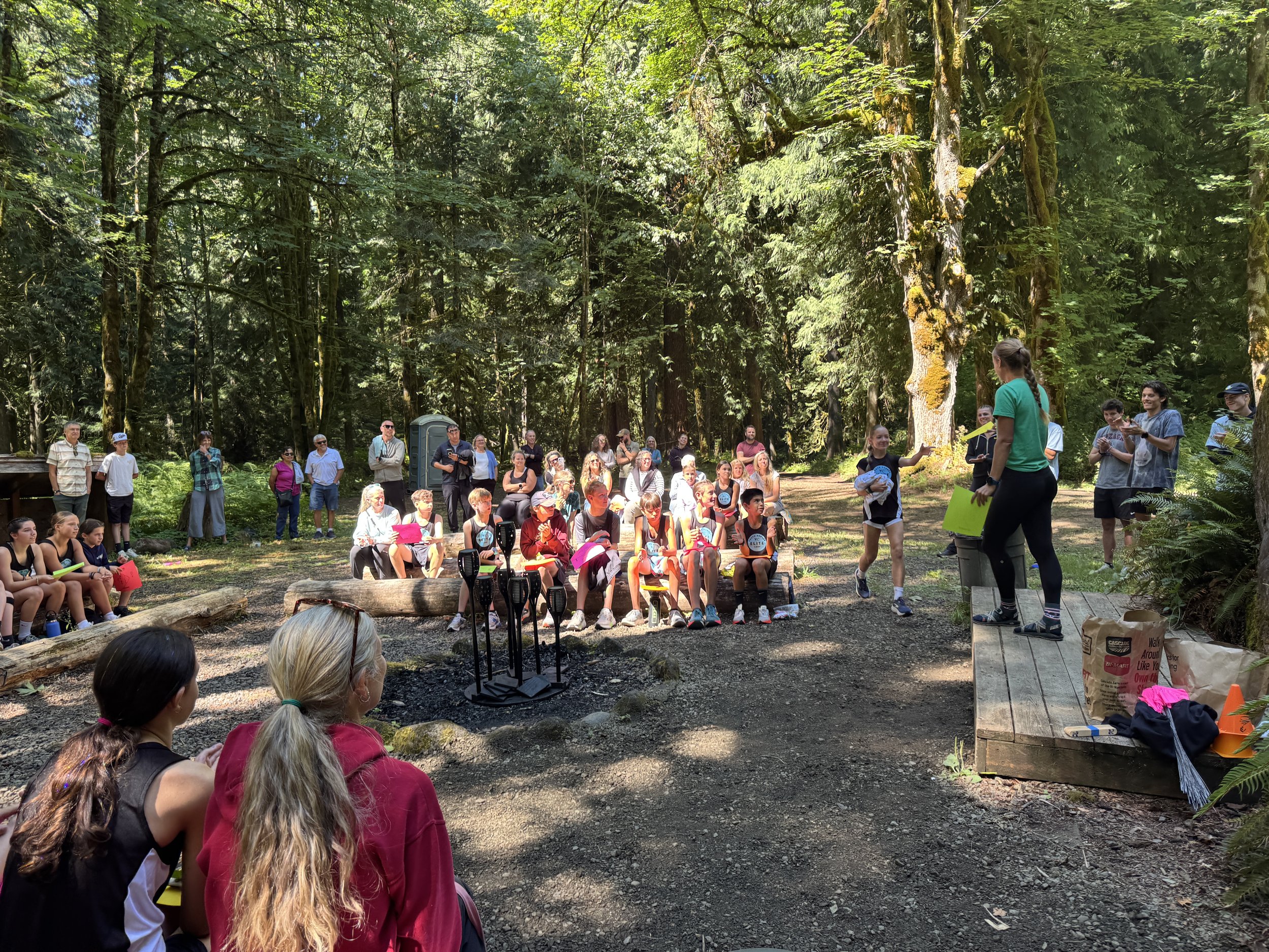 Group of children and adults sitting and standing in a forest clearing, listening to a woman giving a presentation or talk.