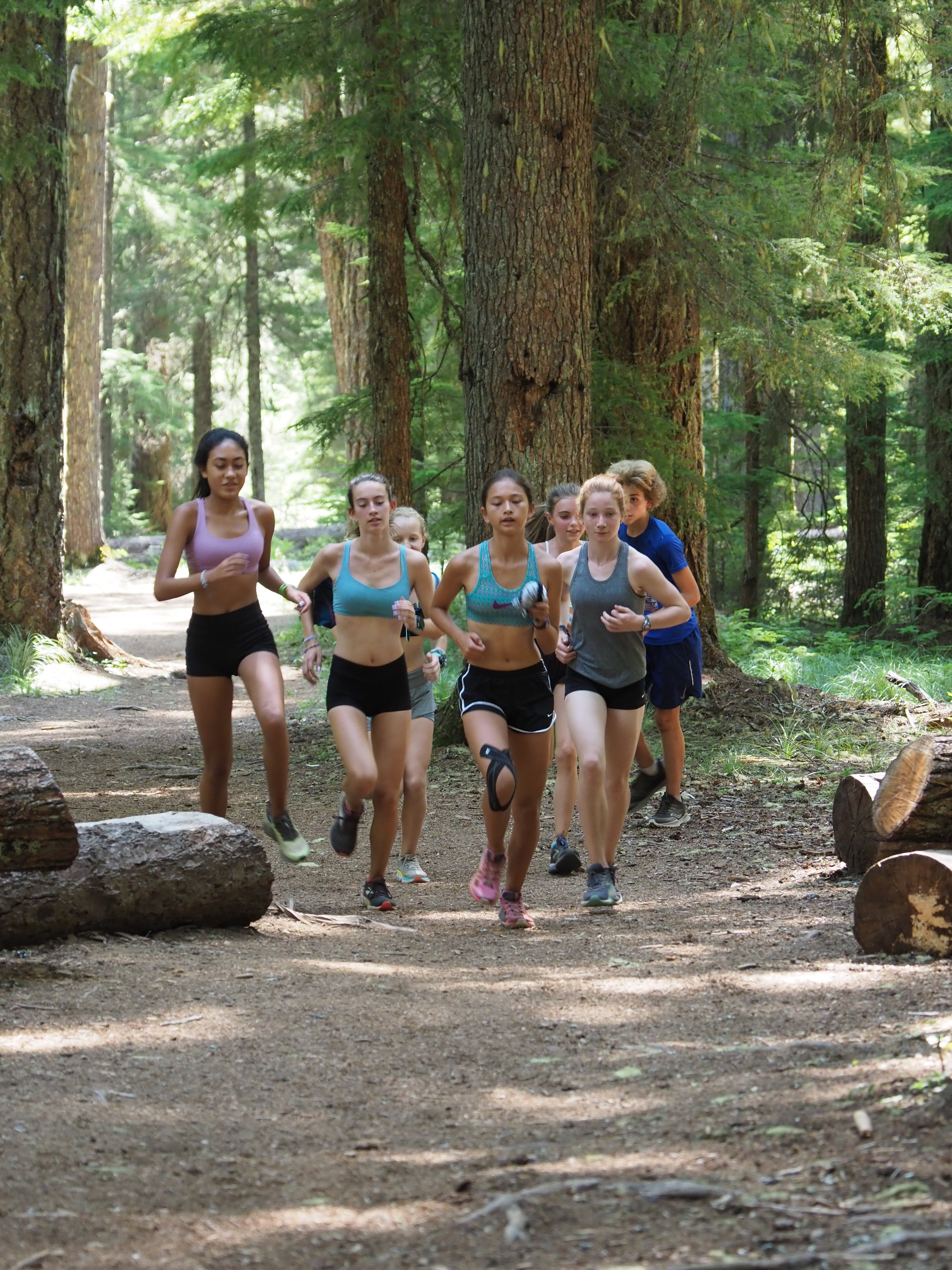 Group of young people running together on a forest trail surrounded by tall trees and greenery.