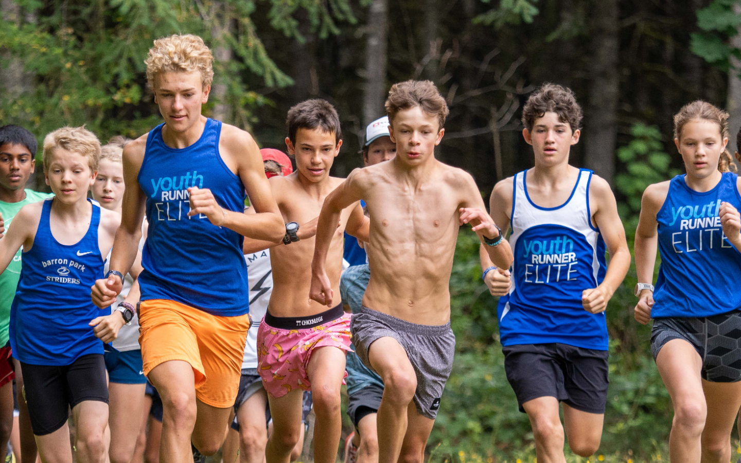 A group of young boys running in a cross-country race through a wooded area, some wearing blue 'Youth Runner Elite' shirts