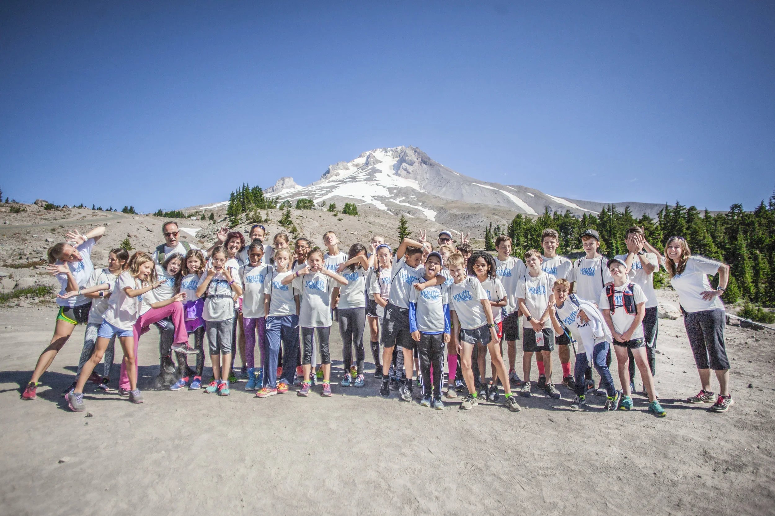 Group of children and adults in outdoor clothing, hiking in front of a snow-capped mountain under a clear blue sky.
