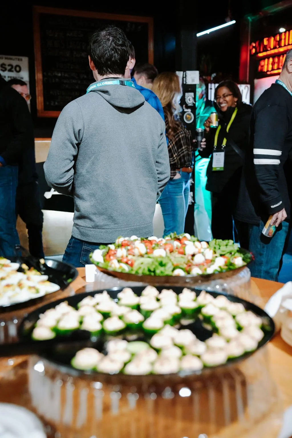 A group of people at a social event in a dimly lit venue with food tables in the foreground and people mingling in the background.