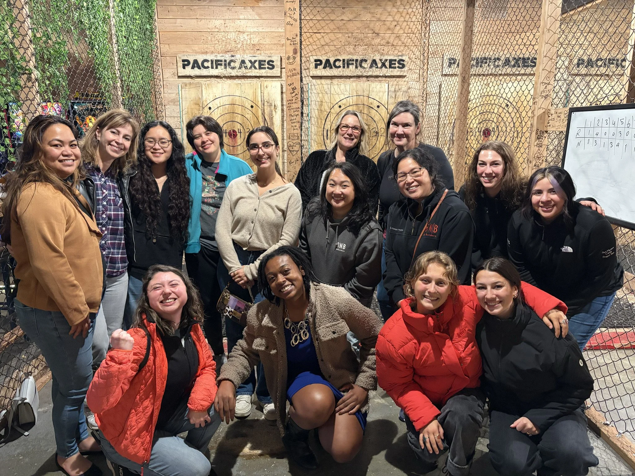 A group of 15 women posing together at an axe-throwing venue, with wooden walls, a target range, and a whiteboard in the background.