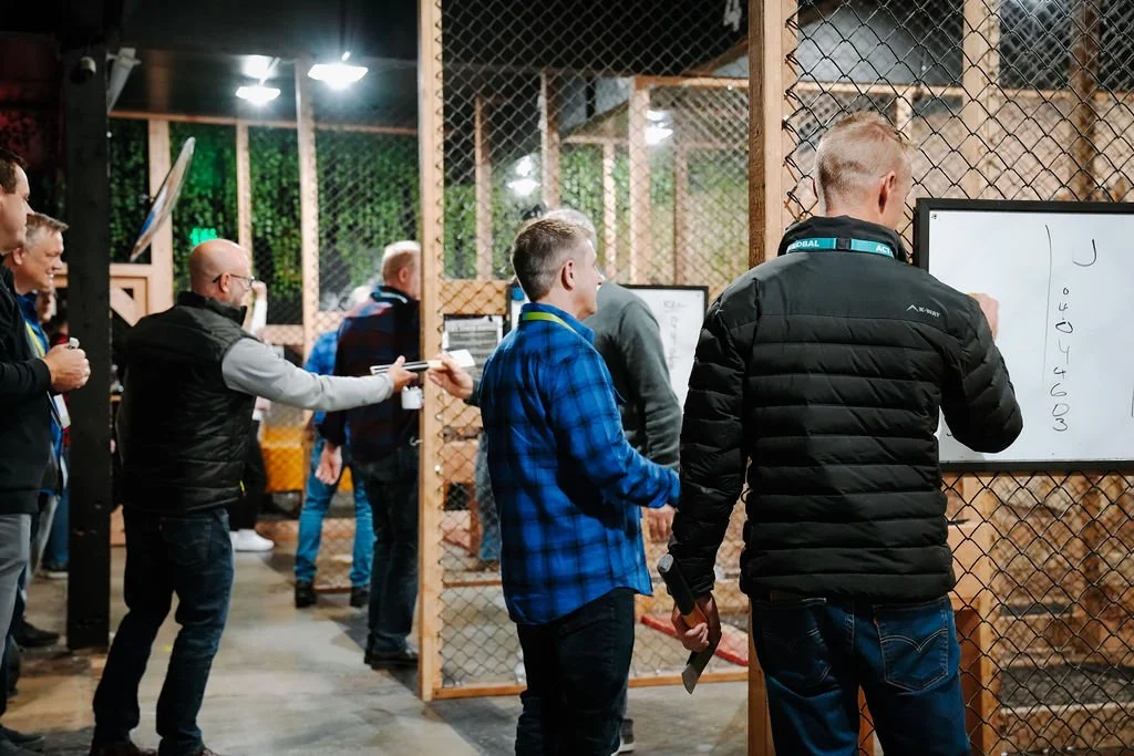 People standing in line at a virtual reality gaming center, with a young man in a blue plaid shirt and an older man in a black jacket writing on a whiteboard.