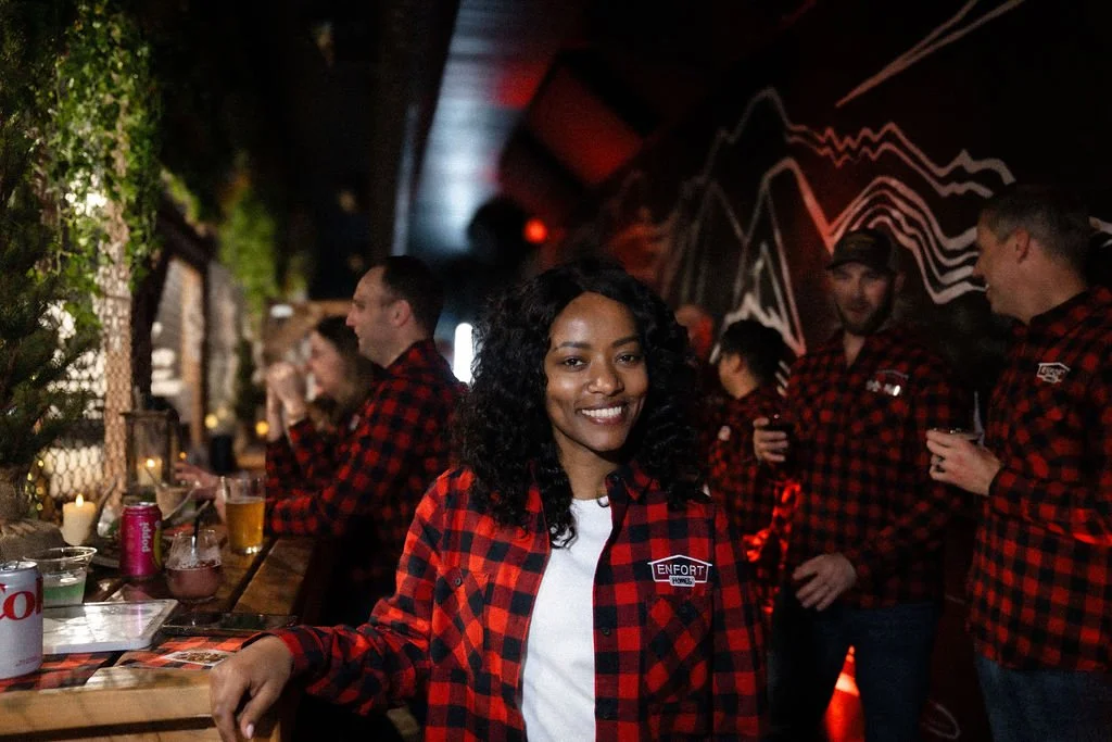Group of people wearing matching red and black plaid shirts at bar or pub, smiling and socializing, with a woman in the foreground.