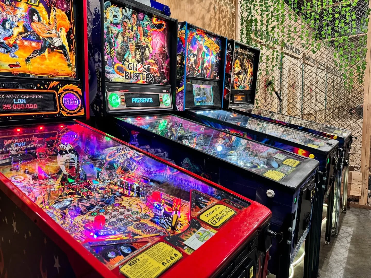 Multiple pinball machines lined up, featuring colorful artwork of rock bands and pop culture icons, with some displaying flashing lights.