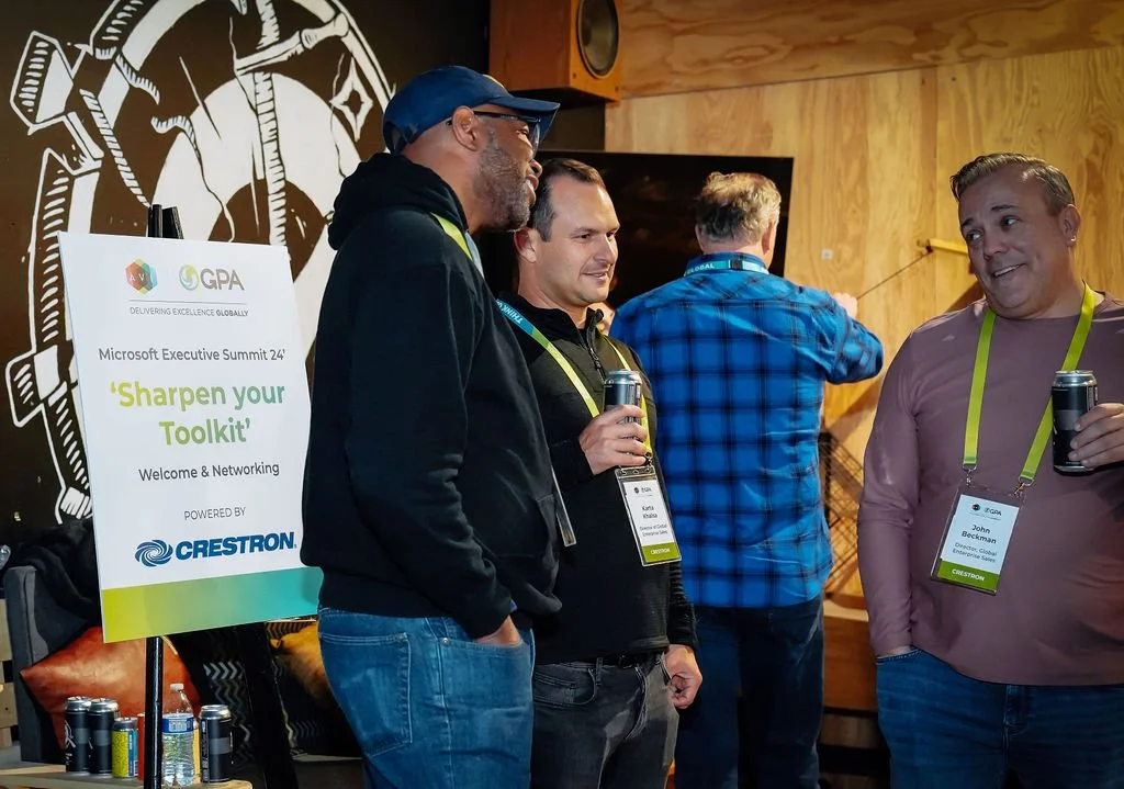 Three men standing and talking at the Microsoft Executive Summit event, with a poster that reads 'Sharpen your Toolkit' in the foreground, and a fourth person in a blue plaid shirt in the background.