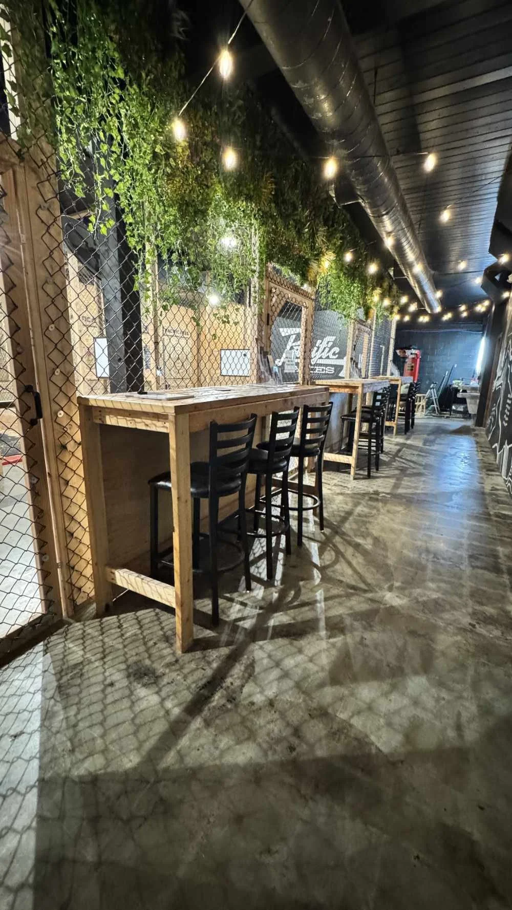 Indoor bar or restaurant with a wooden high table and black chairs, string lights, greenery on the wall, and a polished concrete floor.