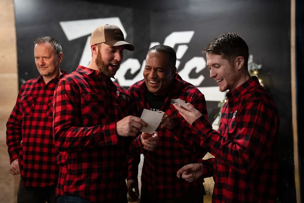 Four men in red and black plaid shirts smiling and looking at a piece of paper and a phone, standing in front of a black wall with white logo.