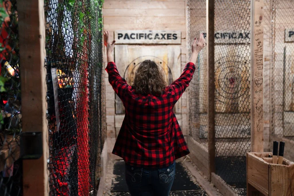 A woman with curly hair wearing a red and black checkered shirt standing in an outdoor axe-throwing area, holding an axe in each hand, with targets on wooden walls in front of her.