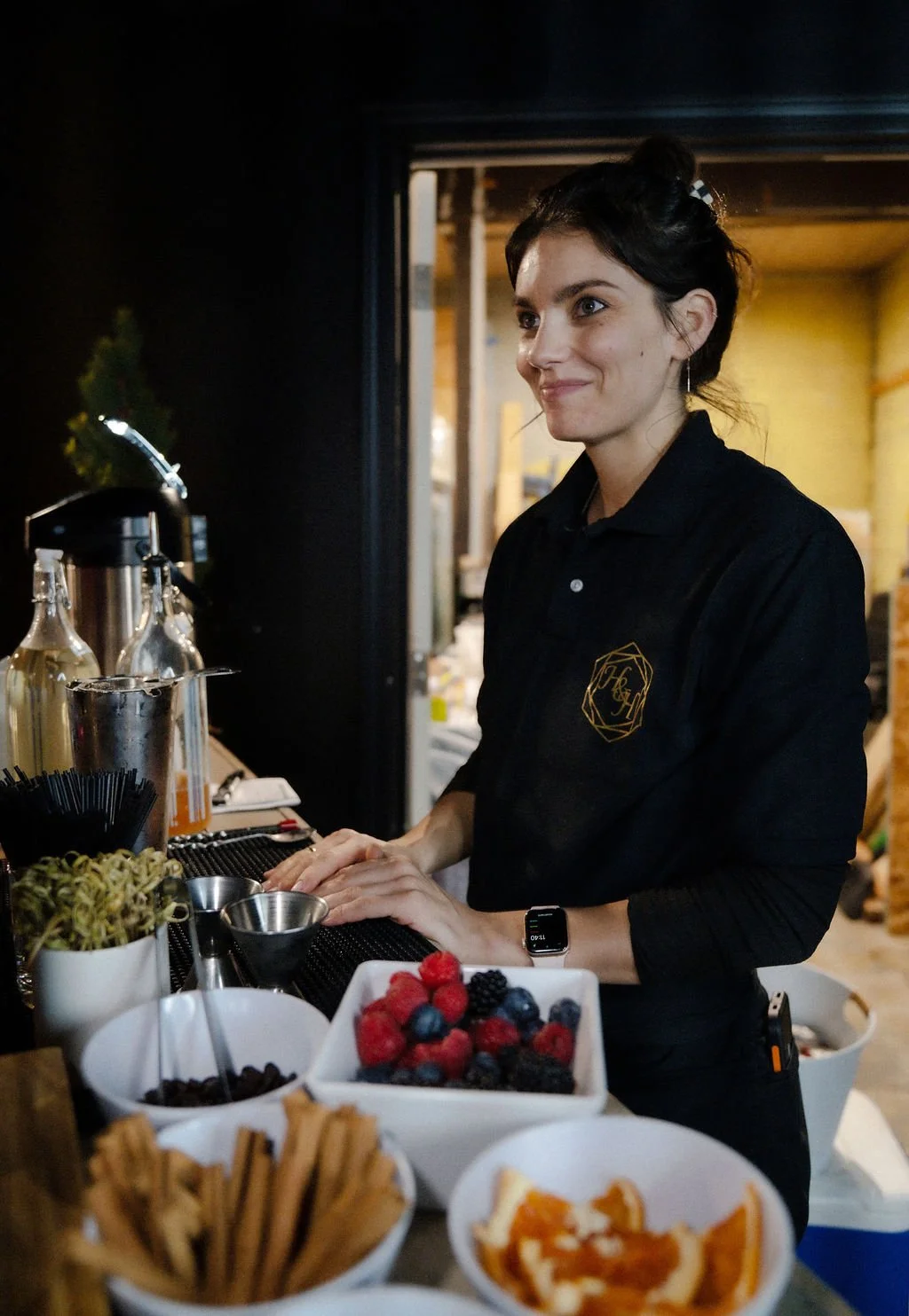 A woman in a black shirt stands behind a bar, preparing drinks with various berries, fruit, and bar tools on the counter.
