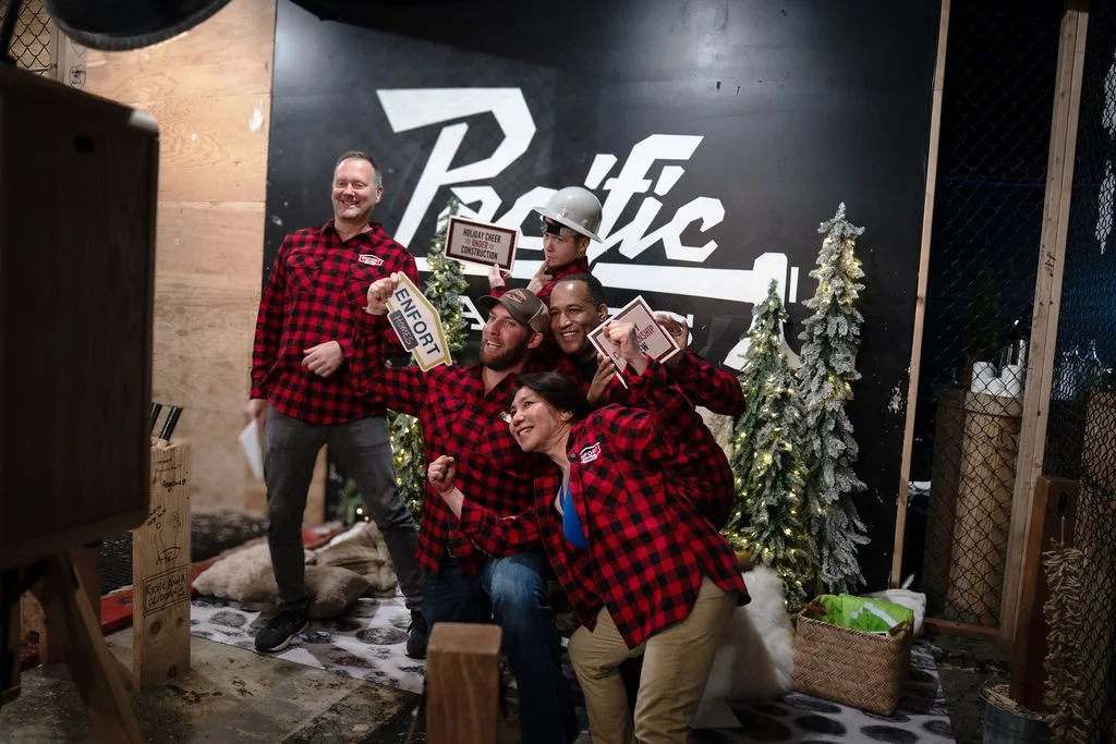Group of five people wearing red and black plaid shirts celebrating indoors with Christmas trees and snow decorations behind them.