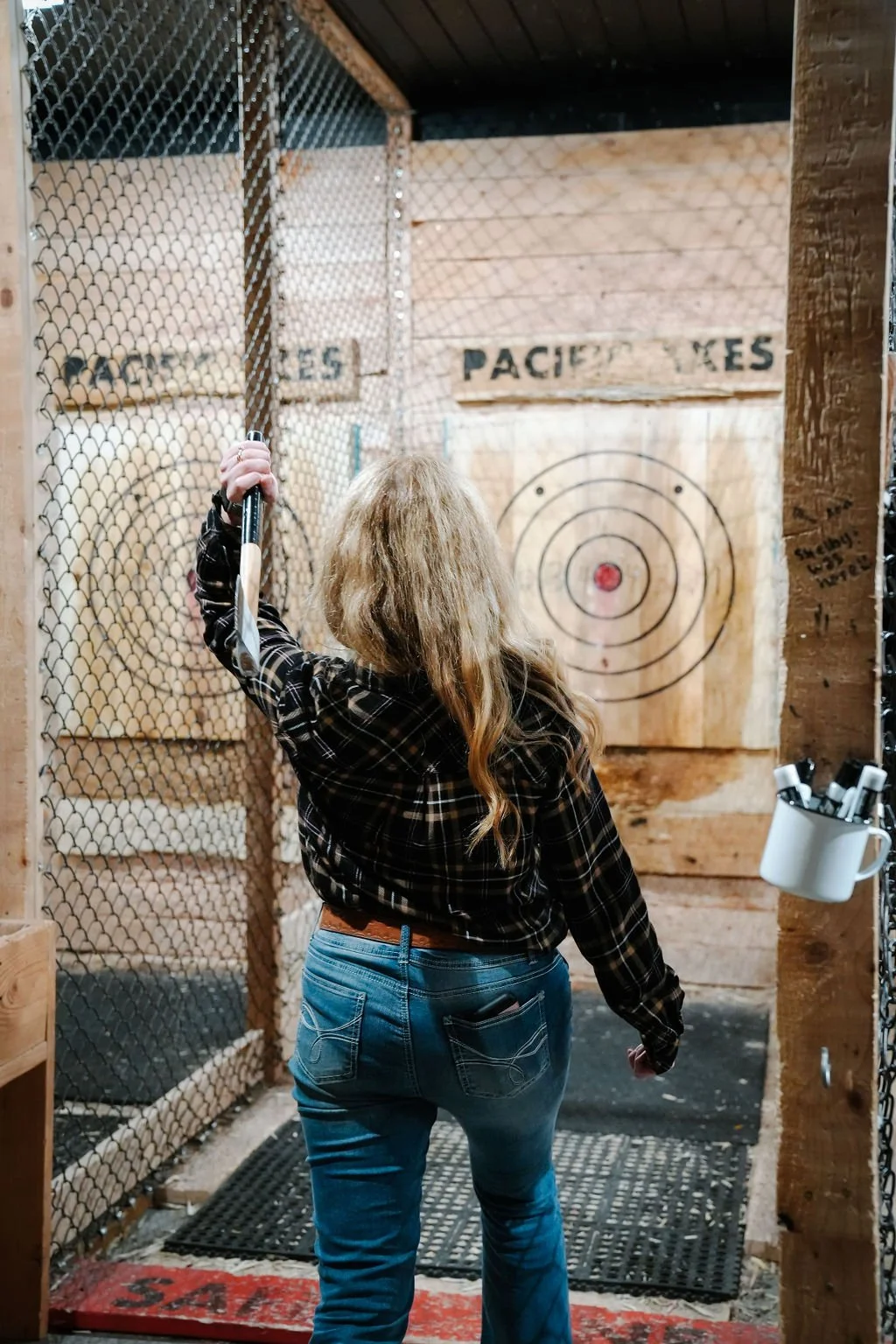 Woman with long blonde hair wearing a black plaid shirt and jeans, holding an axe, about to throw it at an indoor axe-throwing target area with wooden walls, a bullseye, and a chain-link fence.