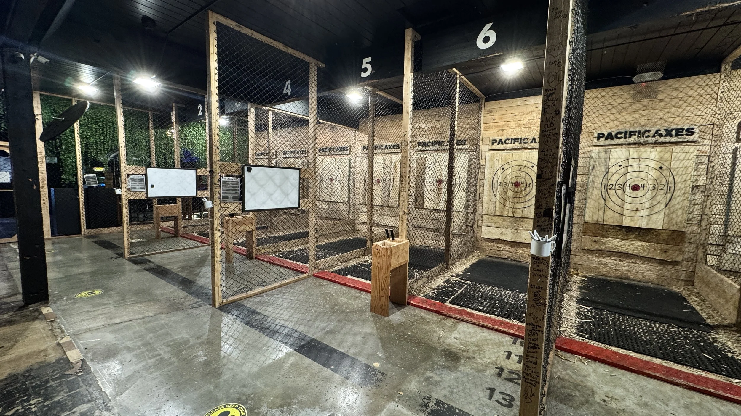 Indoor axe throwing lanes with six designated targets, separated by wooden partitions and black mats, with a wooden container holding axe throwing tools, and whiteboards and scoreboard signs on the fencing.