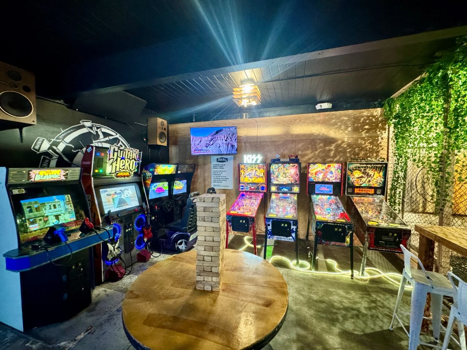 A gaming arcade with multiple pinball machines and arcade cabinets, a large TV on the wall, and a wooden table in the foreground. The room has dim lighting with decorative greenery on the right side.
