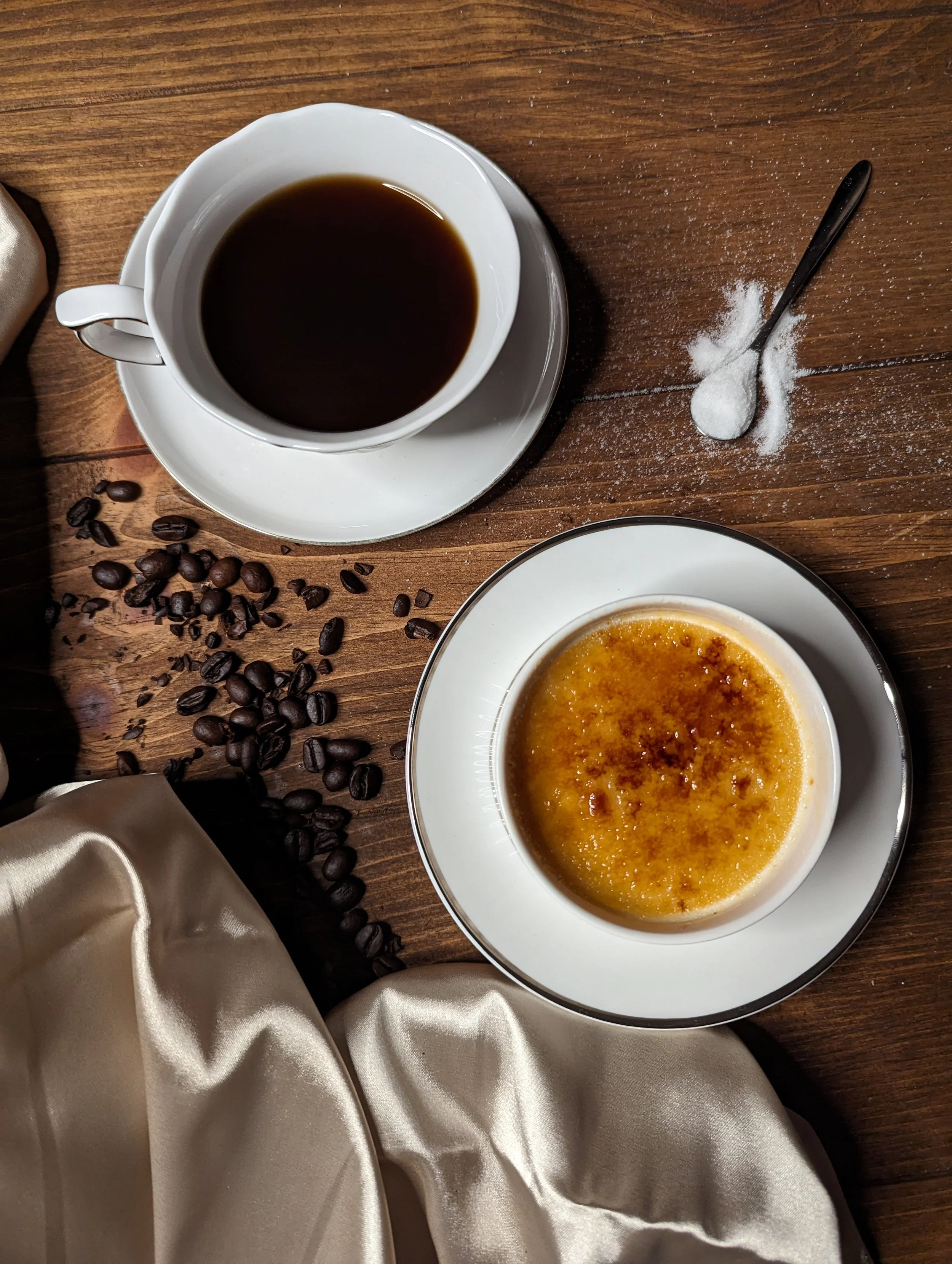 A cup of black coffee, a dish of creme brulee, scattered coffee beans, and a spoonful of sugar on a wooden table with a silk cloth.