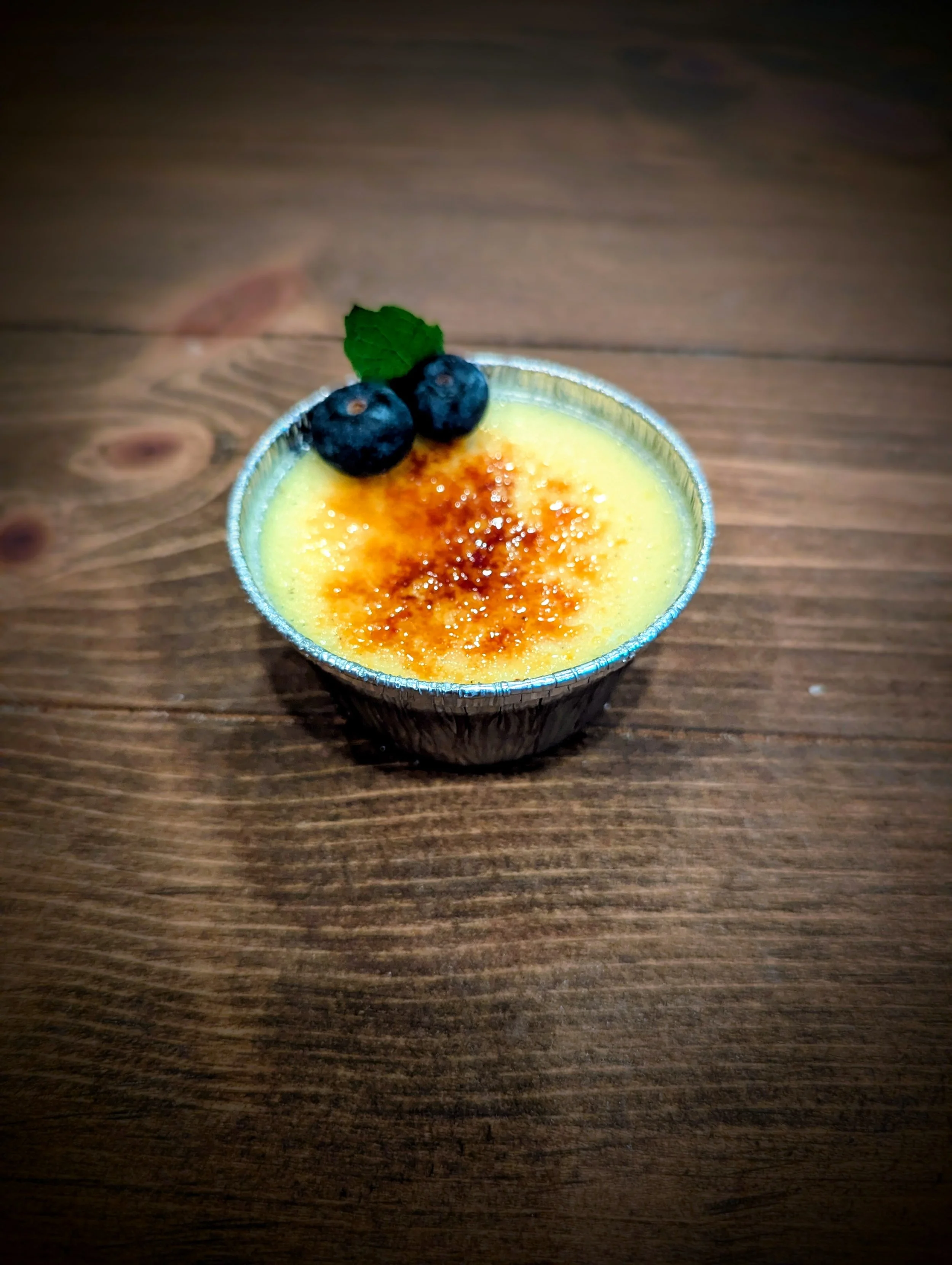 Creme brulee topped with blueberries and a mint leaf, in a small metal cup on a wooden surface.