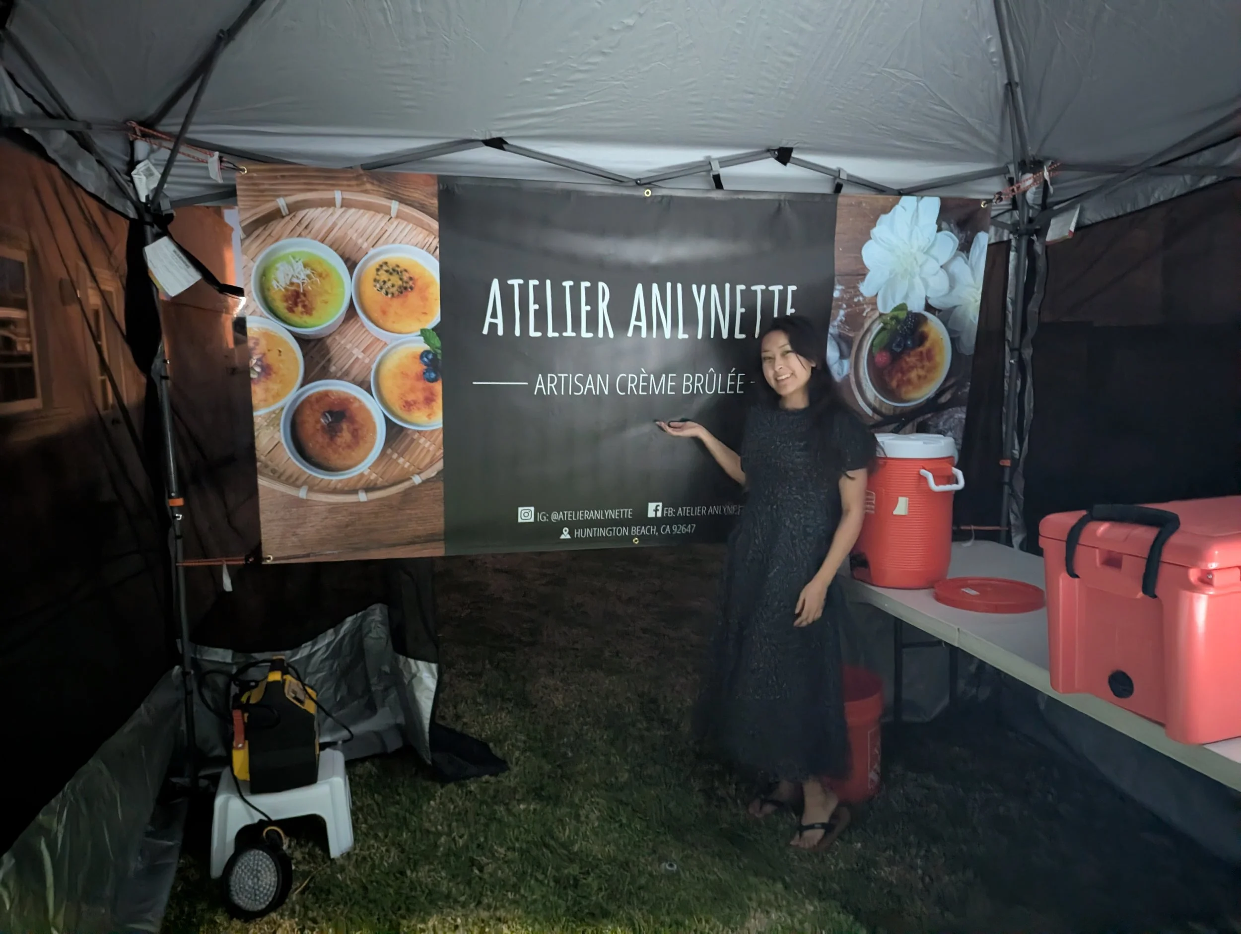 Woman standing inside a tent at a food stall, smiling and pointing to a sign that reads 'Atelier Anlynette, Artisan Creme Brulee'. The stall has a banner displaying pictures of creme brulee desserts, and there are coolers and containers on a table to