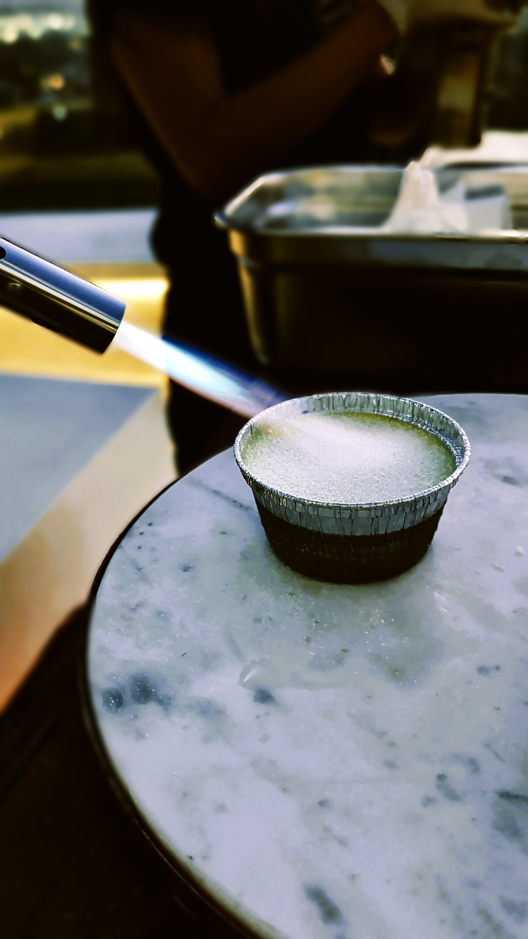 A small black and silver foil container of Crème brûlée on a round table, with a person in the background working with a blowtorch and a metal tray.