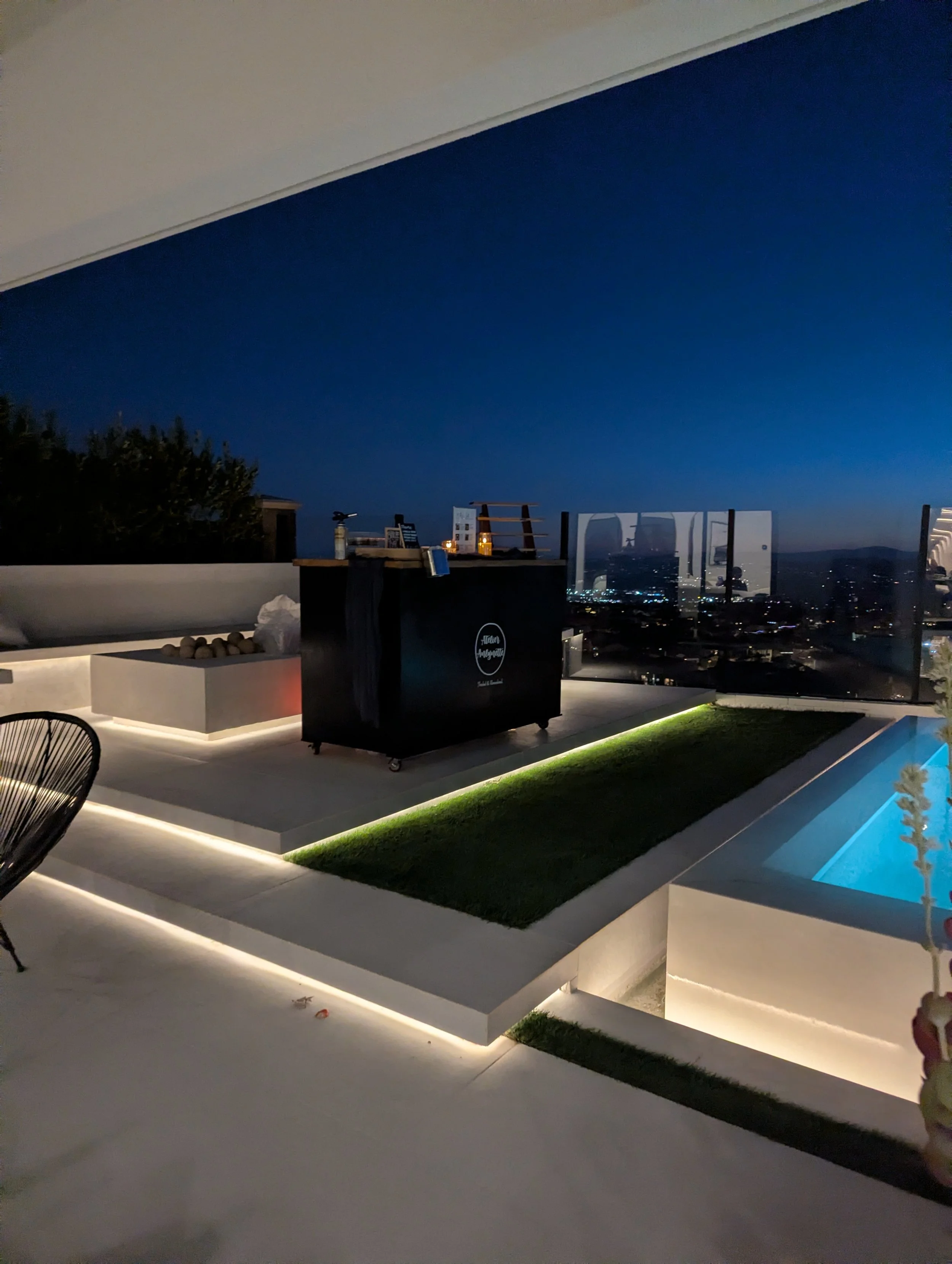 Nighttime rooftop patio with a view of the city skyline, outdoor seating, a pool illuminated with blue light, and a bar with bottles on top.