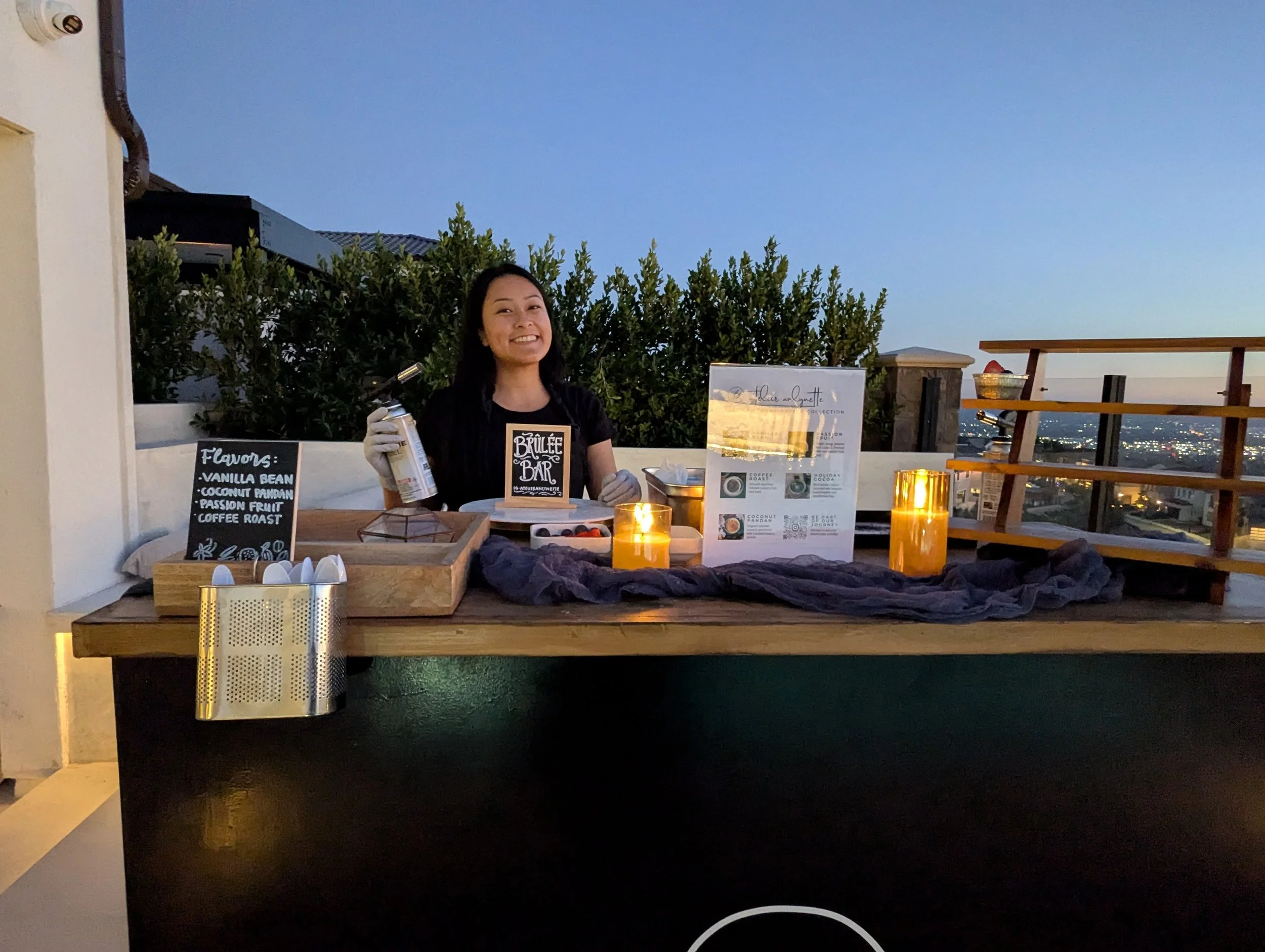 A woman standing behind a wooden table on a rooftop at dusk, smiling, with candles and a menu display, selling flavored coffee drinks.