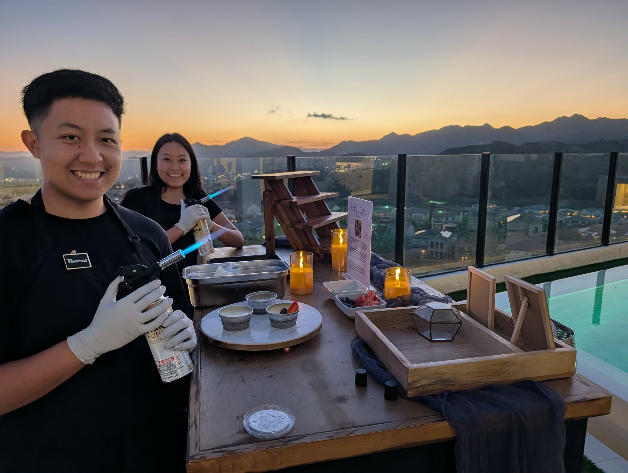 Two smiling people with syringes, on a rooftop at sunset with a city view, preparing food for a culinary experience.