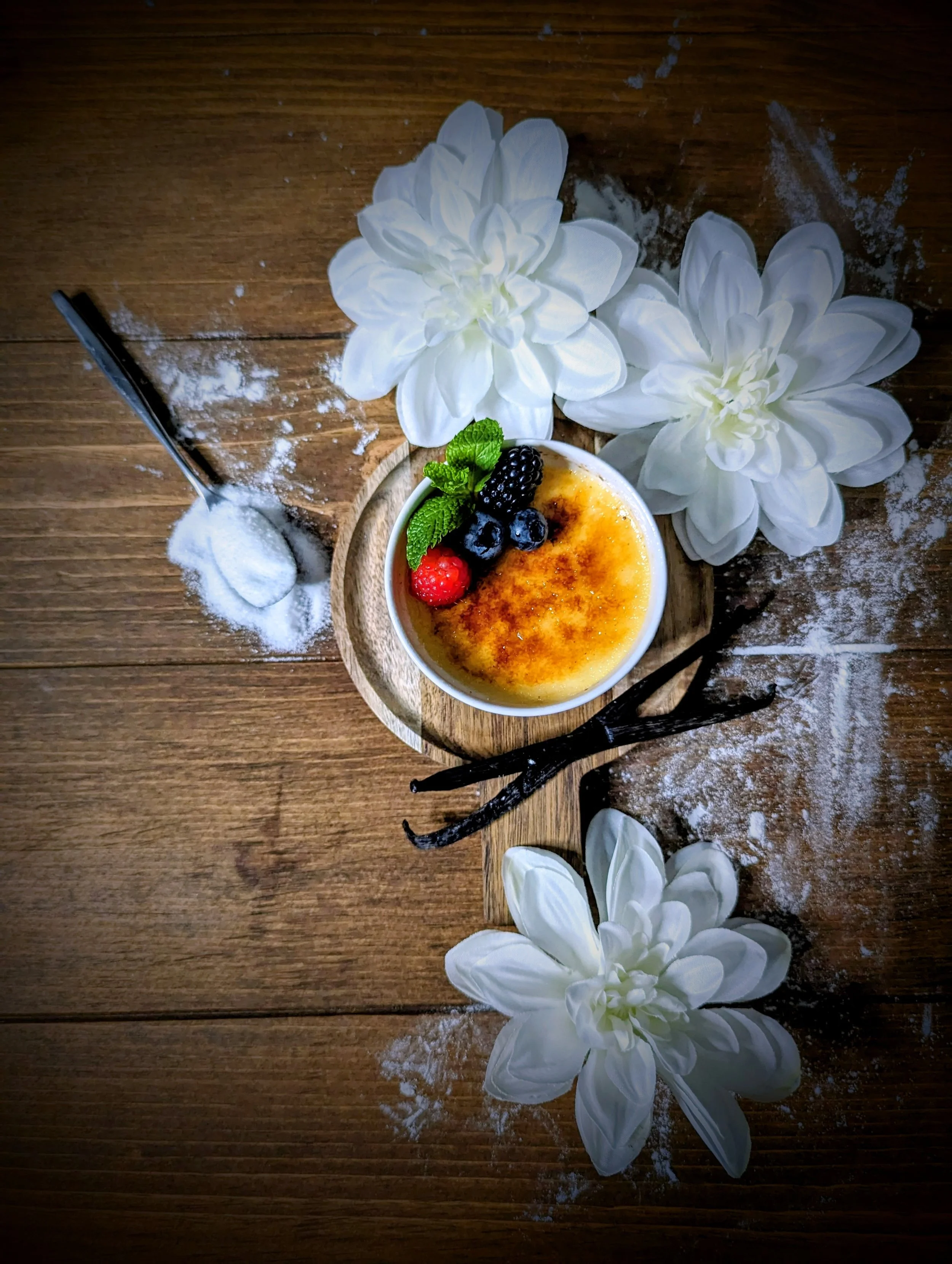 A dessert of crème brûlée garnished with berries and mint leaves, surrounded by white flowers and vanilla beans on a wooden surface with powdered sugar.