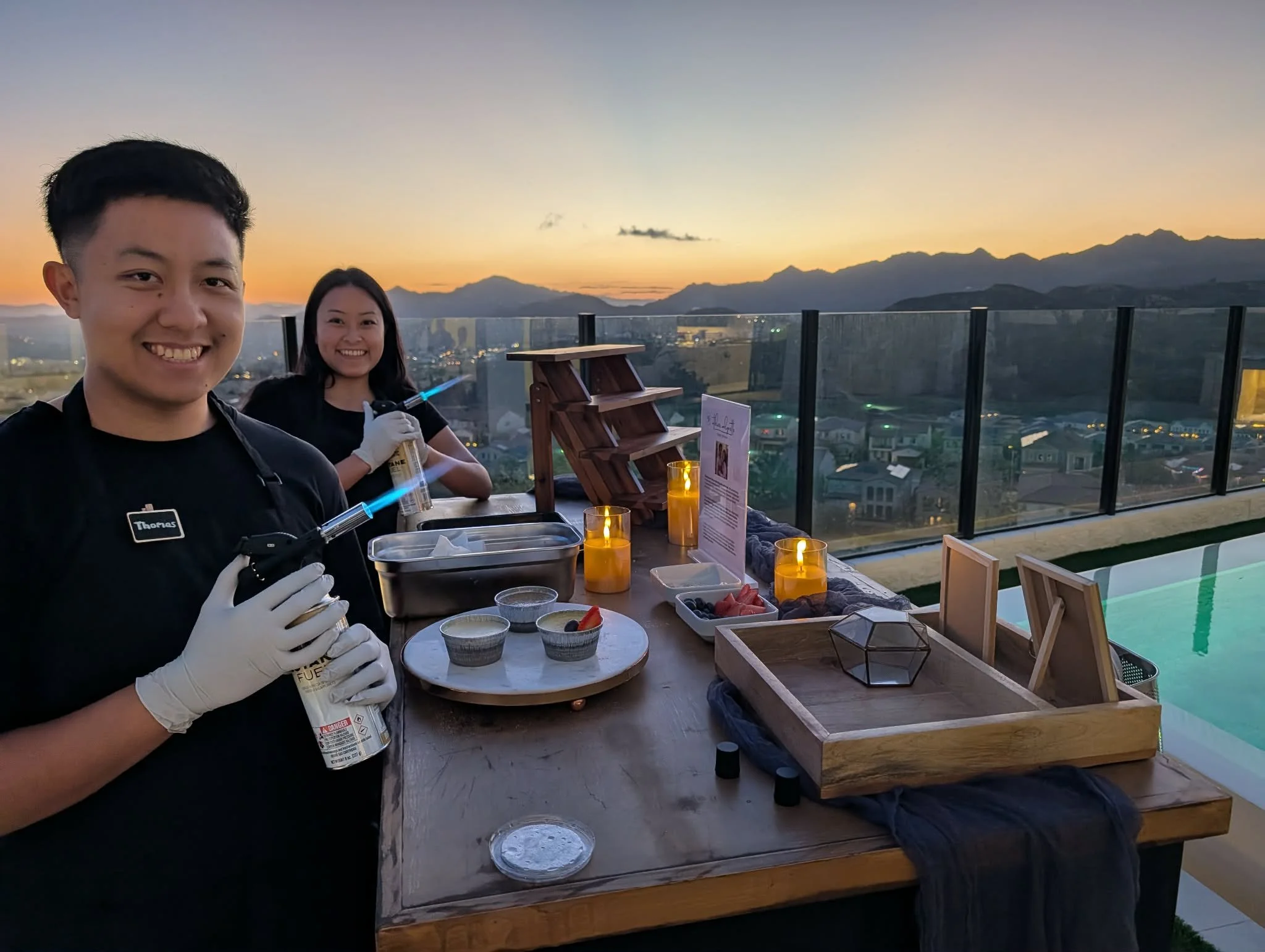Two people preparing sushi at a rooftop dinner table during sunset with a cityscape and mountains in the background.