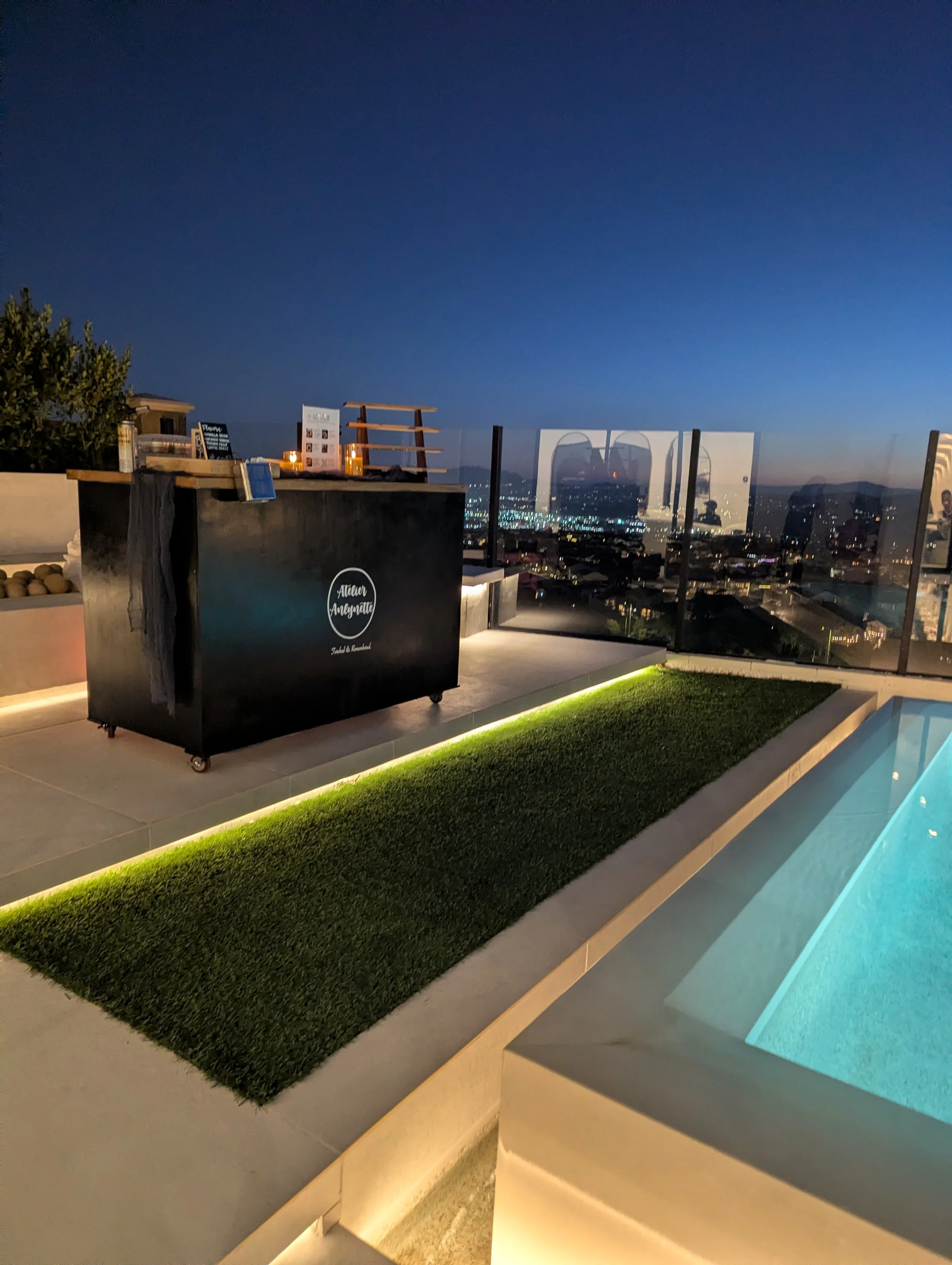 Rooftop bar area at night with city skyline in the background, a pool with blue water, artificial grass, and illuminated lighting under the railings.